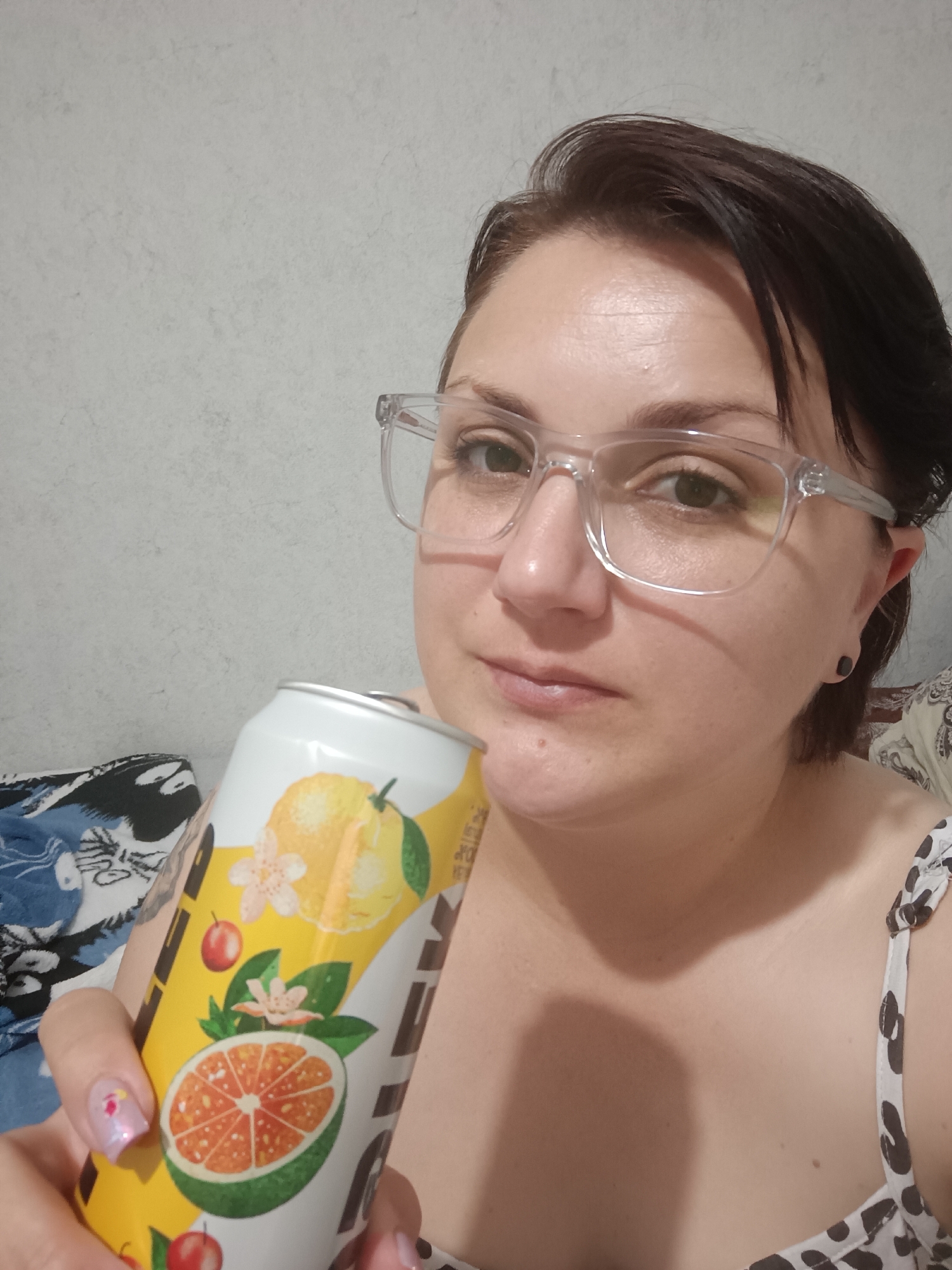 A close-up, selfie-style shot features a woman in her late 20s or early 30s holding a can of beverage. She has fair skin, dark brown hair styled short, and is wearing clear-rimmed glasses. Her eyes are green and she is looking slightly to her left. The can she is holding is predominantly white with a yellow design featuring illustrations of citrus fruits, flowers, and berries. The woman's right hand is visible, showcasing her painted fingernails. She is wearing a sleeveless top with a black and white patterned design. The background is a plain, textured wall. The image appears to be taken indoors, possibly in a residential setting. There is no discernible information in the image to indicate the location in Krasnodar, Russia.