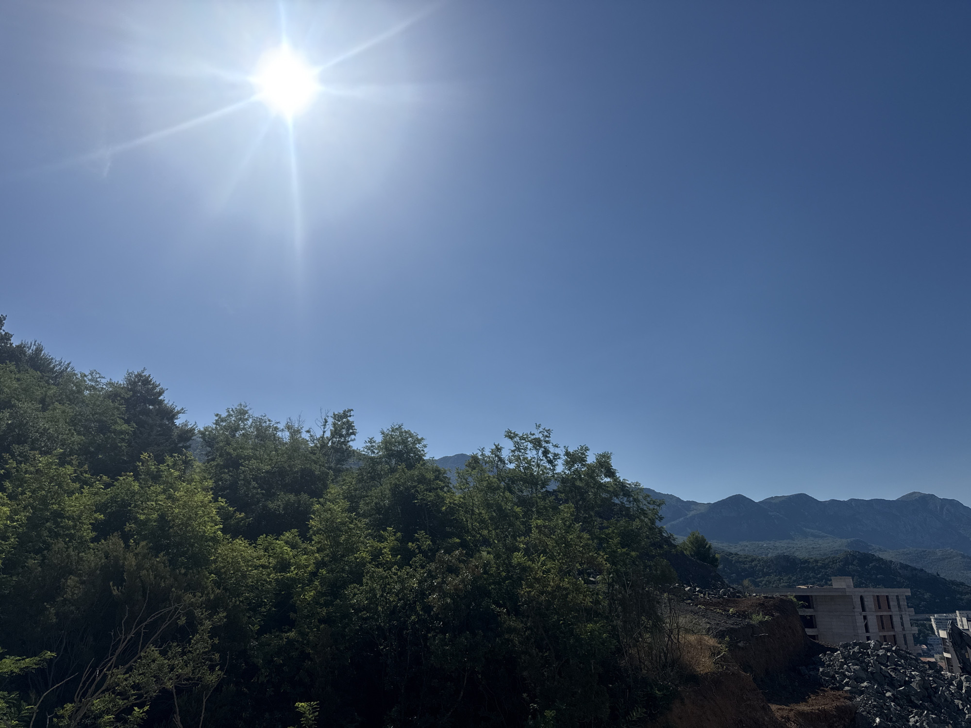 The original image depicts a bright, sunny day with a clear blue sky dominating the upper portion of the frame. The sun, a brilliant white orb with visible rays, is positioned in the upper left quadrant, casting a strong glare. Below the sky, a lush green hillside covered in dense foliage and trees extends across the middle and left of the image. The foliage appears thick and healthy, with varying shades of green indicating different types of trees and plants.
On the right side of the image, the hillside transitions into an area of construction. A multi-story building, appearing to be under construction, is visible with its concrete structure exposed. Piles of rocks and debris are scattered around the construction site, suggesting active work or recent excavation. Further in the background, a range of majestic mountains with undulating peaks stretches across the horizon, their forms softened by distance and atmospheric haze. The overall scene suggests a mountainous coastal region under clear, sunny weather.
Specifically referencing the city of Boreti, Montenegro, this image appears to capture a view from a hillside overlooking the coastline or a bay. Boreti is known for its picturesque setting with a mix of natural beauty and developing infrastructure. The presence of construction on the hillside could indicate ongoing development within or near the city. The combination of dense vegetation, mountains, and the construction site aligns with the typical landscape features found in this region of Montenegro, which is known for its beautiful Adriatic coastline and mountainous hinterland.