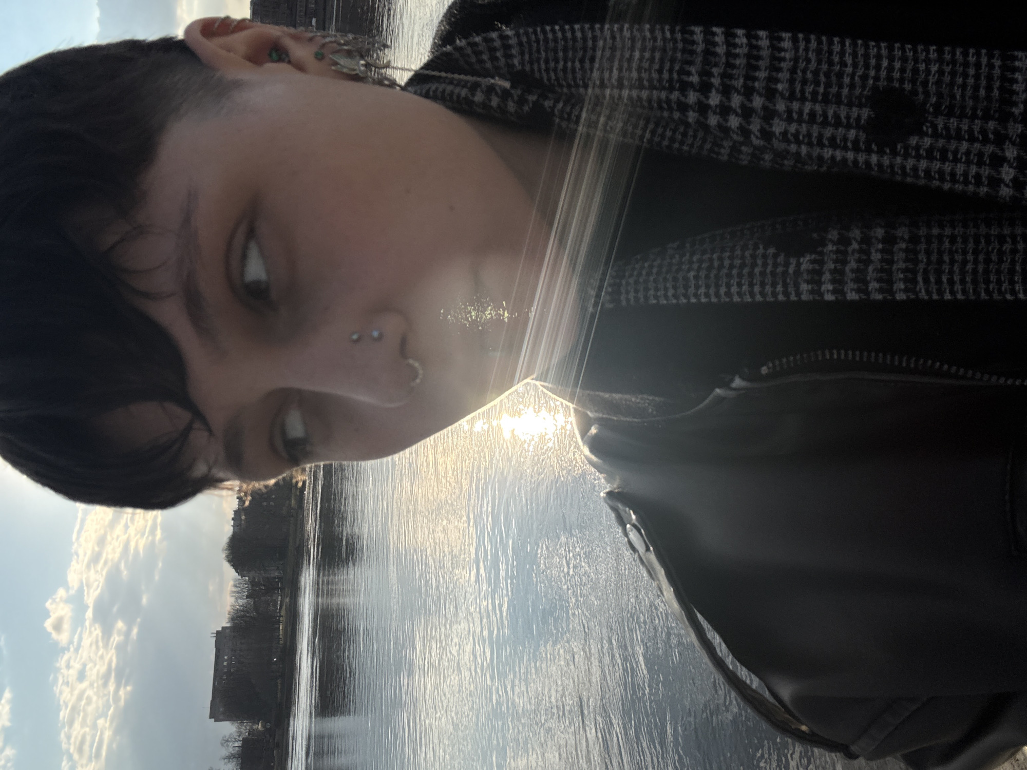 A low-angle, slightly rotated, close-up shot features a person in the foreground with a river and buildings visible in the background. The person, likely female, has dark hair with a short, shaved section and a longer fringe. Multiple piercings are visible on the nose and ear, including a stud on the nostril and several ear piercings with ornate jewelry. The person is wearing a black leather jacket and a black-and-white houndstooth-patterned scarf. The face is partly obscured by sunlight reflecting off the river. The river appears to be the Neva River, and the buildings on the opposite bank suggest this location is in Saint Petersburg, Russia. The sky is partly cloudy with blue patches. The water reflects the sunlight, creating bright, shimmering patterns.
