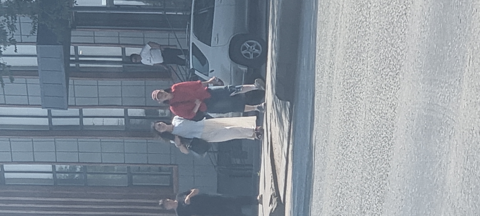 In Aktobe, Kazakhstan, on a paved street, two women walk side-by-side. The woman on the left wears a white top and long white pants, carrying a black handbag. The woman on the right is dressed in a red long-sleeved shirt, dark shorts, and a red baseball cap. A person in a white shirt and dark pants stands on a ledge in front of a building. A silver SUV is parked behind the women. The building features large, grid-like windows and appears to be modern architecture.