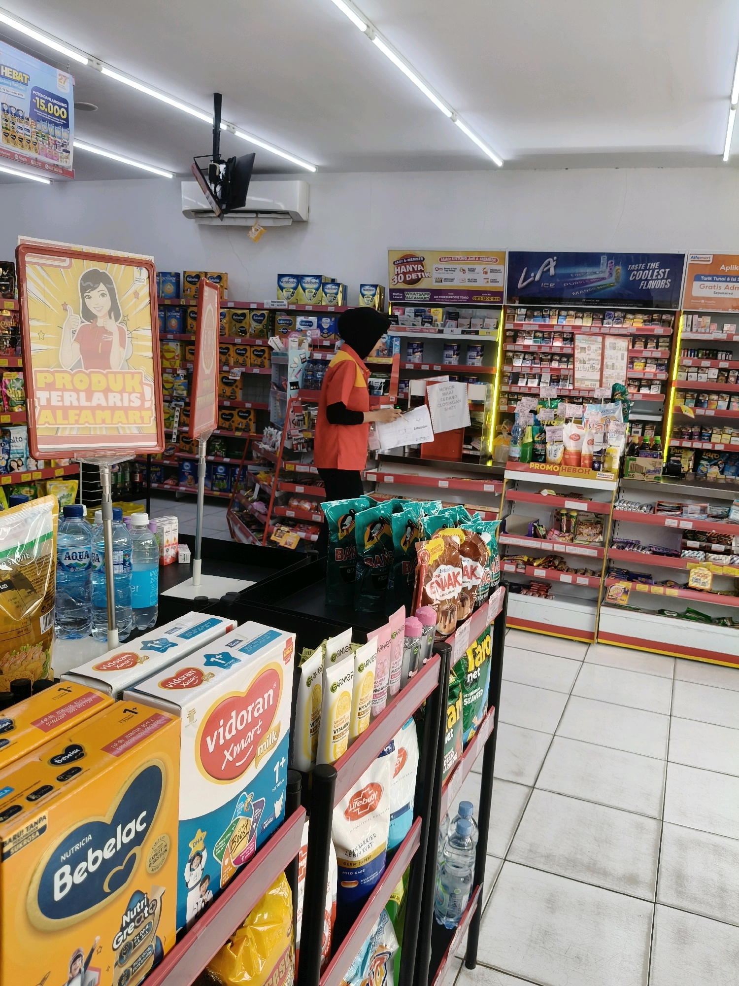 The image depicts the interior of an Alfamart convenience store, a common retail chain in Indonesia, specifically located in South Tangerang. A female store employee, wearing an orange and black uniform and a headscarf, stands behind a counter, appearing to be processing paperwork or interacting with a customer off-camera. Various products are displayed on shelves and in display racks, including bottled water, baby formula (Bebelac and Vidoran Xmart milk), and packaged snacks. Promotional signage, such as a banner advertising "PRODUK TERLARIS ALFAMART" (Alfamart's Bestselling Products), is visible. The store environment is well-lit with fluorescent lighting, and the floor is tiled. An air conditioning unit is mounted high on the wall. The scene captures a typical retail setting during operating hours.