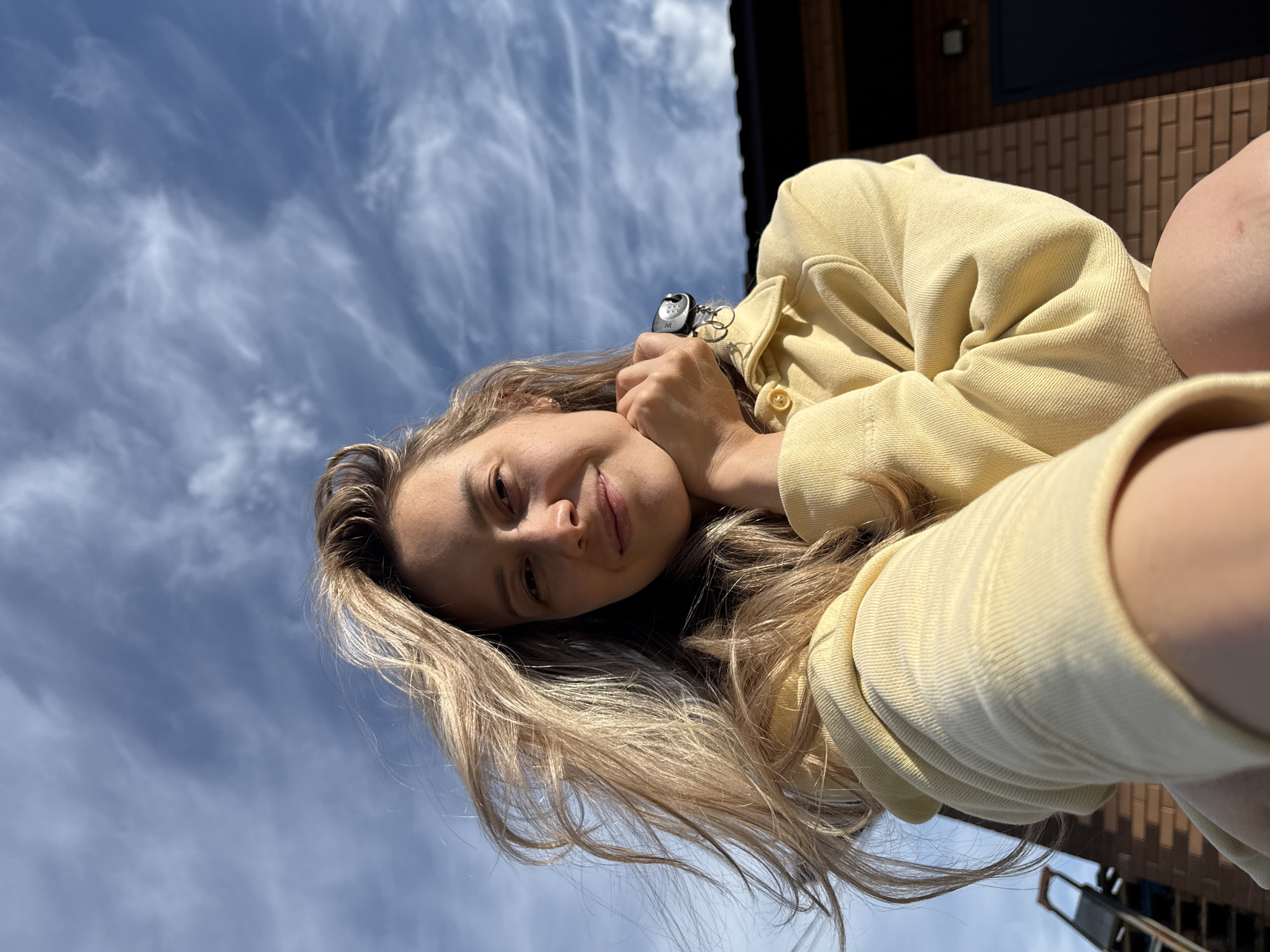 A young woman with long blonde hair is seen from a low angle against a backdrop of a cloudy sky and a building facade. She is wearing a yellow jacket. She is holding a set of keys near her face and smiling slightly. The building's facade is made of brown bricks, and the location is in Chaikovsky, Russia.