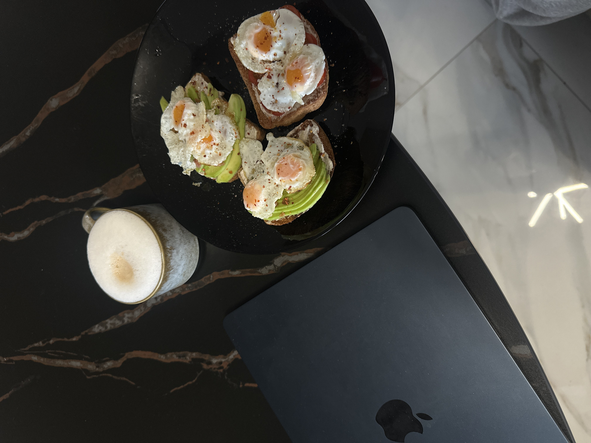 An overhead shot captures a breakfast or brunch scene set on a modern, dual-toned tabletop in Sofia, Bulgaria. The table surface on the left is dark, possibly black or dark brown, and features prominent reddish-brown and white marble-like veining. To the right, a contrasting lighter, white or light gray surface with subtle gray veining creates a distinct boundary, suggesting either two adjoining surfaces or a large table with varied patterns.
Central to the image is a black, round plate holding three pieces of toast. Each toast is generously topped with sliced avocado and two sunny-side-up fried eggs, which are visibly sprinkled with red pepper flakes. A creamy spread is discernible beneath the avocado and eggs on all pieces. One of the toasts also features slices of red tomato beneath its egg toppings.
To the lower left of the plate, a light-colored ceramic mug with a subtle gold rim contains a frothy, light brown beverage, likely coffee or a latte, with small bubbles on its surface. Adjacent to the plate and mug, resting on the dark tabletop, is a closed dark gray or black laptop, clearly identifiable as a MacBook due to the distinct Apple logo embossed on its lid. The overall arrangement suggests a calm and relaxed setting for a daytime meal, possibly combined with work or leisure activities. No people are visible in the frame.