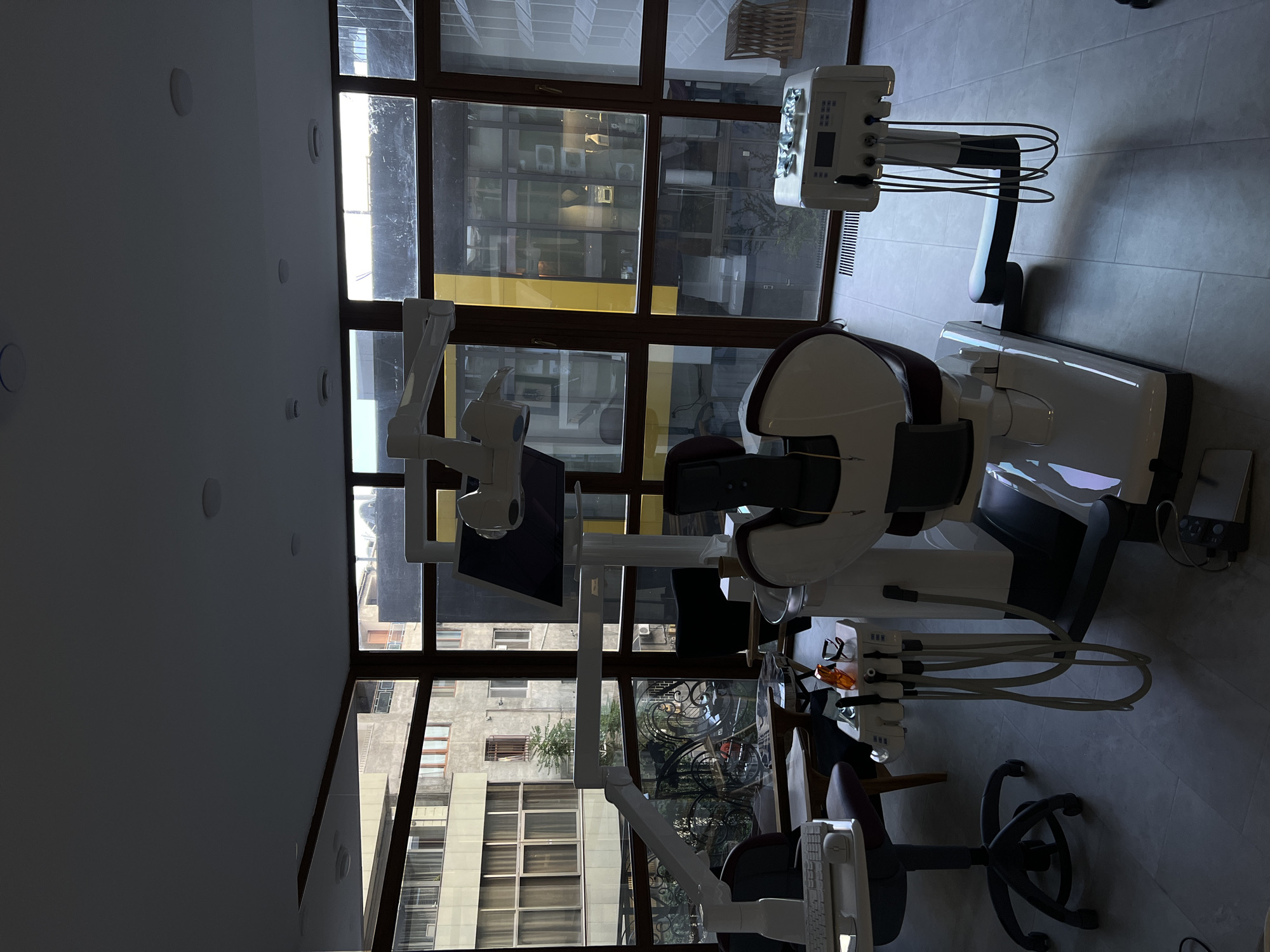 The image shows a dental office in Yerevan, Armenia. The focus is on a modern dental chair unit positioned in front of a large window with a dark wood frame. The chair is white and brown, with accompanying equipment like a monitor, overhead light, and dental tools. The window offers a view of surrounding buildings, revealing the urban setting. The flooring appears to be tiled. No people are visible in the scene.