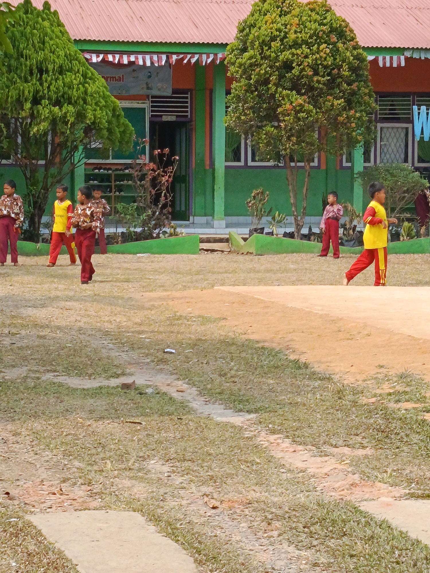 The image depicts a sunny outdoor scene at what appears to be a school in Megang Sakti IV, Indonesia. In the foreground, a dirt and grass field is visible, with a light-colored path cutting through it. Several children, dressed in school uniforms, are scattered across the scene. Two children in the left midground wear patterned shirts and maroon pants, while another child to their right wears a yellow shirt and red pants. A child in the right midground is dressed in a yellow shirt and red pants, with red stripes down the legs. Behind the children, a building with a red corrugated roof and green walls stands. The building has several windows with patterned glass and a visible entrance. Decorative red and white banners are strung across the top of the building's facade. Lush green trees with full foliage frame the building on the left and right sides. The overall environment suggests a casual school day activity.