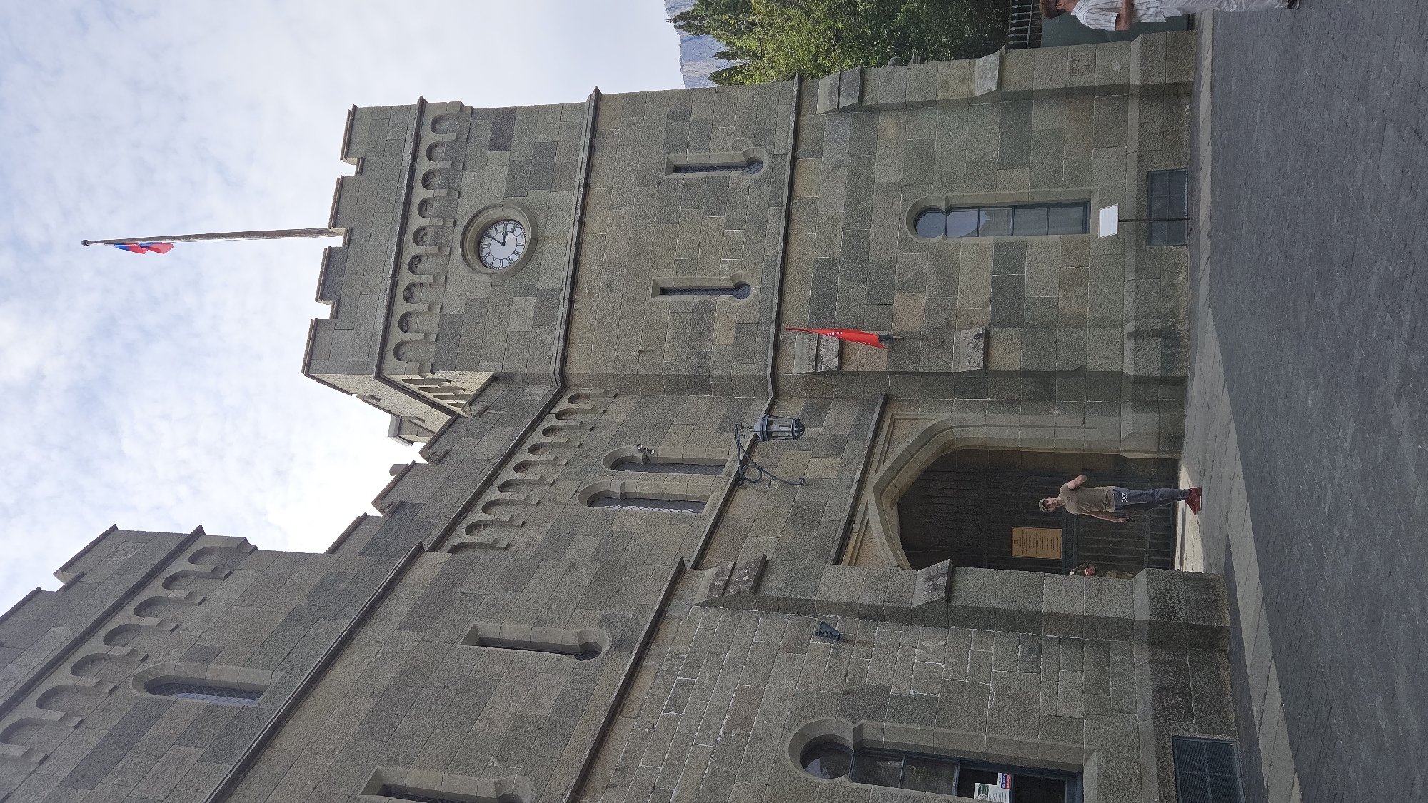 The image shows the Vorontsov Palace in Alupka, Crimea. The palace is made of grey stone and features a clock tower with a working clock. A Russian flag flies from the tower. There are several people present, including a man standing in the entrance archway. The pavement is visible in the foreground. The sky is partly cloudy.