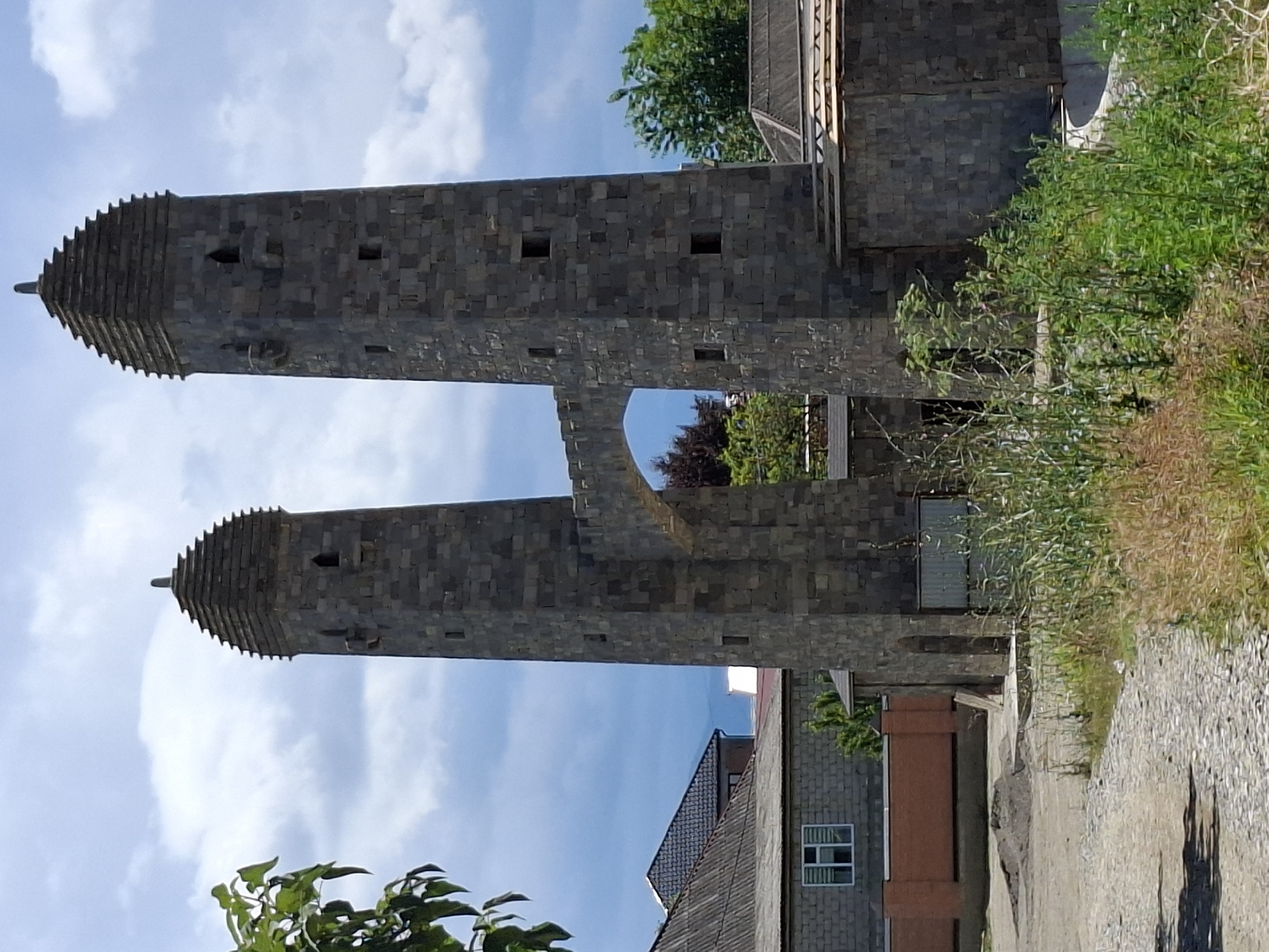 The image displays two identical stone towers, connected by an arched walkway. The towers have a tiered, pyramidal roof, topped with a pointed finial. Small rectangular openings are visible on the towers. The towers are constructed from large, dark gray stone blocks. The walkway between the towers is also made of stone and features a decorative, crenelated edge.
In the background, to the left of the towers, is a low-rise building with a grey tiled roof and a brick wall. Further to the left, a tree with green leaves is partially visible. To the right of the towers, another building with a wooden roof structure is present. The foreground is occupied by a dirt path paved with cobblestones, and an area of tall, green vegetation, some of which is dry and brown. The sky is cloudy with patches of blue. The scene appears to be an outdoor architectural site, possibly a historical monument or a newly constructed structure designed to resemble historical architecture. The location is identified as the city of Urus-Martan, Russia.