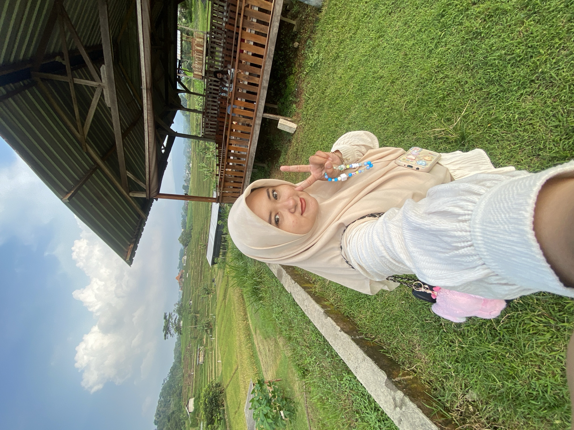 A young woman in a beige hijab is taking a selfie in the city of Penanggungan, Indonesia. She is making a peace sign with her fingers. She is wearing a light-colored top. A beaded bracelet is visible on her wrist. She has a phone with a decorative case placed on her lap. She is sitting on a grassy area. Behind her is a wooden structure with a roof, and beyond that, a terraced rice field landscape is visible under a partly cloudy sky. A concrete structure or border runs along the edge of the grass where she is sitting. A pink fluffy object attached to a black purse chain is visible in the lower right.