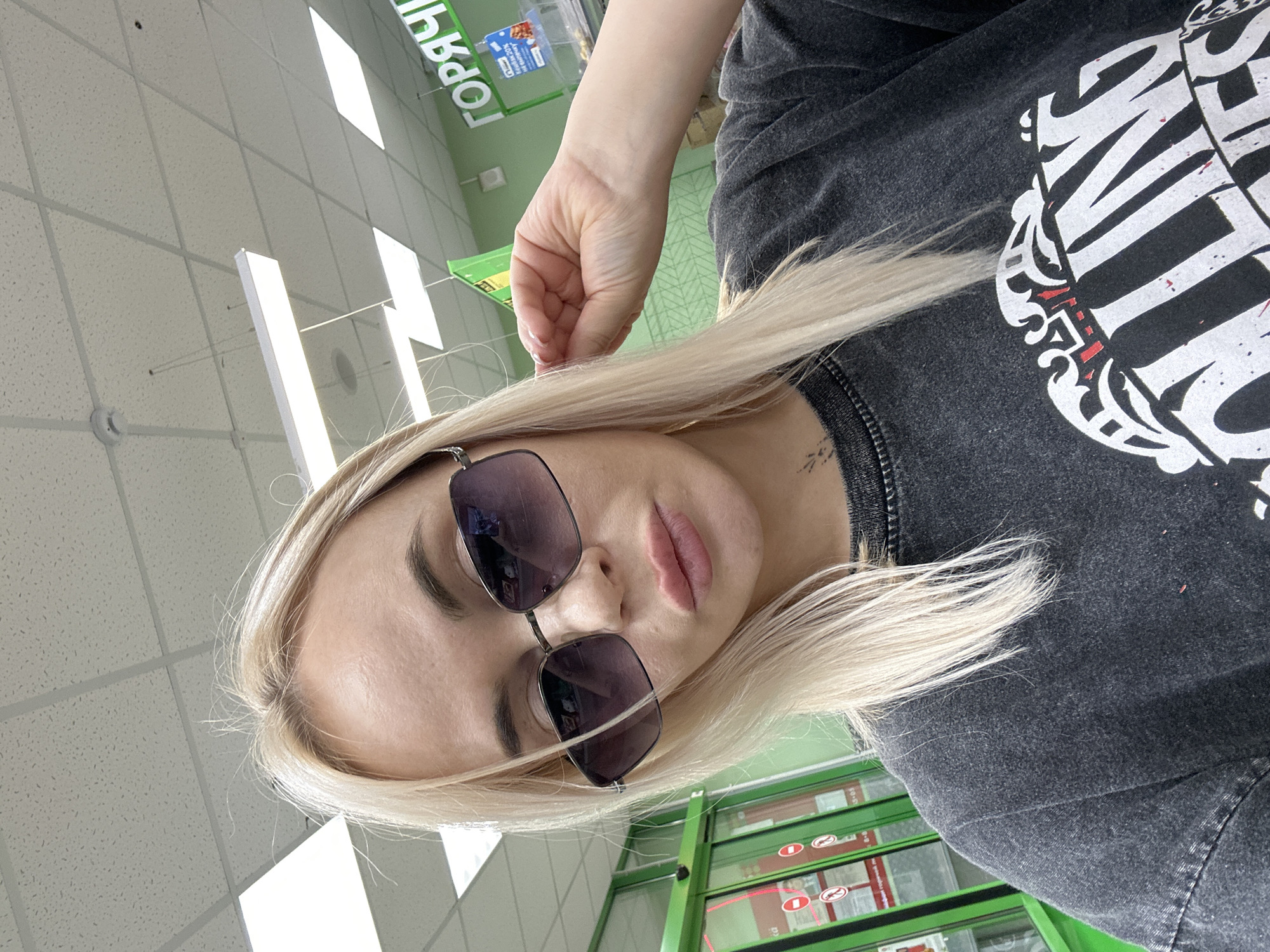 A woman with blonde hair is wearing sunglasses and a dark-colored shirt. She is in a supermarket in Высокий, Russia, based on the Russian text on the signs in the background. The supermarket has a white tile ceiling with fluorescent lights. The woman is looking directly at the camera.