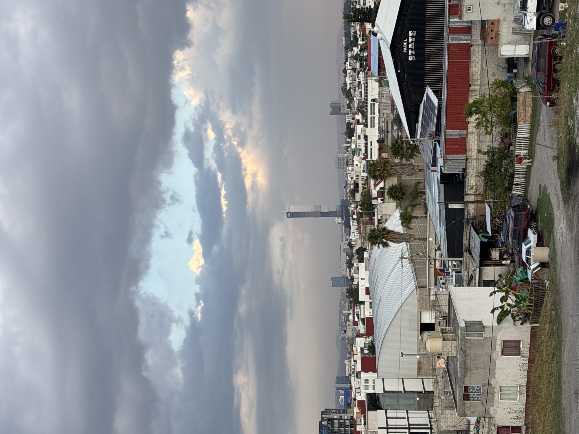 The image depicts a section of San Bernardino Tlaxcalancingo, Mexico, under a cloudy sky. The landscape consists of densely packed buildings, primarily residential, interspersed with commercial structures. There are many residential buildings with white and red rooftops. The built environment shows a mix of architectural styles and building materials. The photograph includes a sports complex building with metal roof, and a 'Padel State' structure visible in the area. Some trees and vegetation can be seen amidst the buildings. In the foreground is some debris including red vehicles and pallets. The skyline is punctuated by taller structures. The overall impression is one of a dense, populated urban area on the edge of the city.