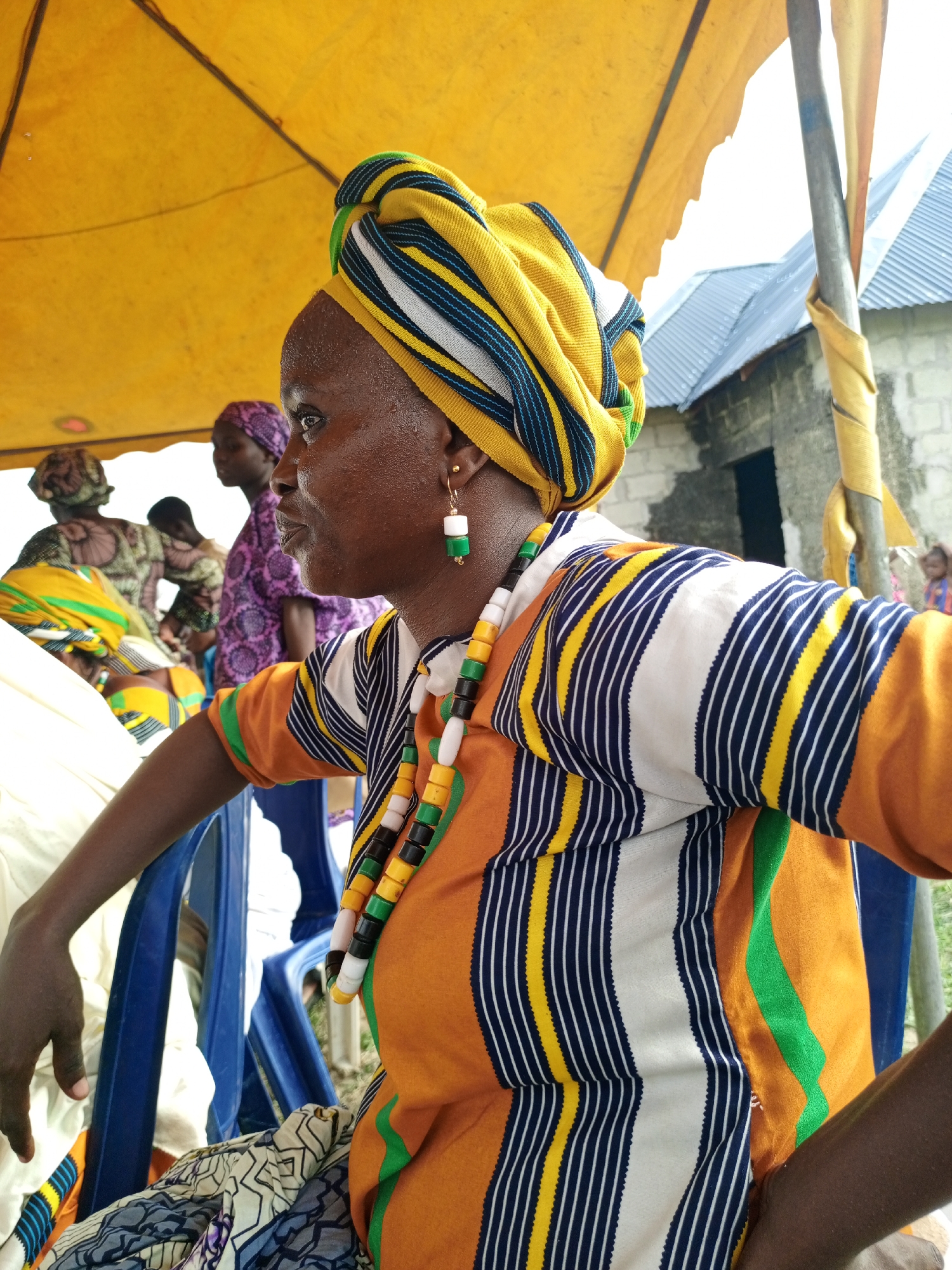 A woman wearing a yellow, blue, and white striped headwrap and a matching striped shirt is sitting in a blue plastic chair under a yellow canopy in Keffi, Nigeria. The shirt has orange and green accents. She is also wearing a beaded necklace and earrings. Several other people are in the background, some wearing patterned clothing. A building with a corrugated metal roof is visible in the background.