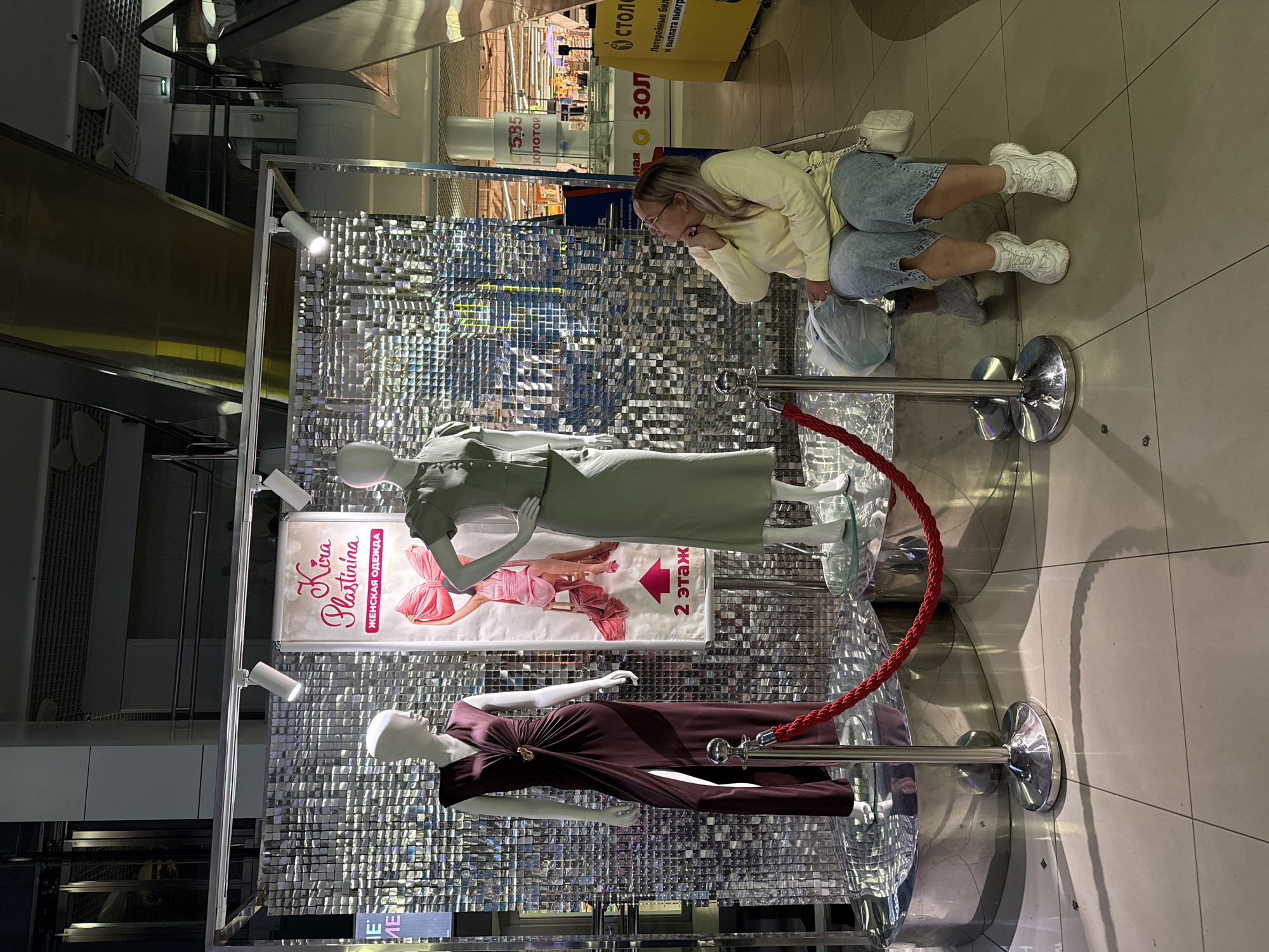 In Ermakovo, Russia, a woman is seated in front of a retail display featuring two mannequins dressed in women's clothing. The display is against a shimmering, sequined backdrop. The mannequin on the left wears a dark, floor-length dress with a high slit and a gold accent. The mannequin on the right wears a light green dress with a collar. A pink banner between the mannequins reads "Kira Plastinina" and "ЖЕНСКАЯ ОДЕЖДА" (Women's Clothing) along with an arrow indicating "2 этаж" (2nd floor). A red rope barrier is in place in front of the display. The woman, wearing a light yellow long-sleeved shirt, denim shorts, and white sneakers, is looking upwards at the display. She has a white purse over her shoulder and is holding a plastic bag. The surrounding environment appears to be a shopping mall, with escalators and other retail signage visible in the background.