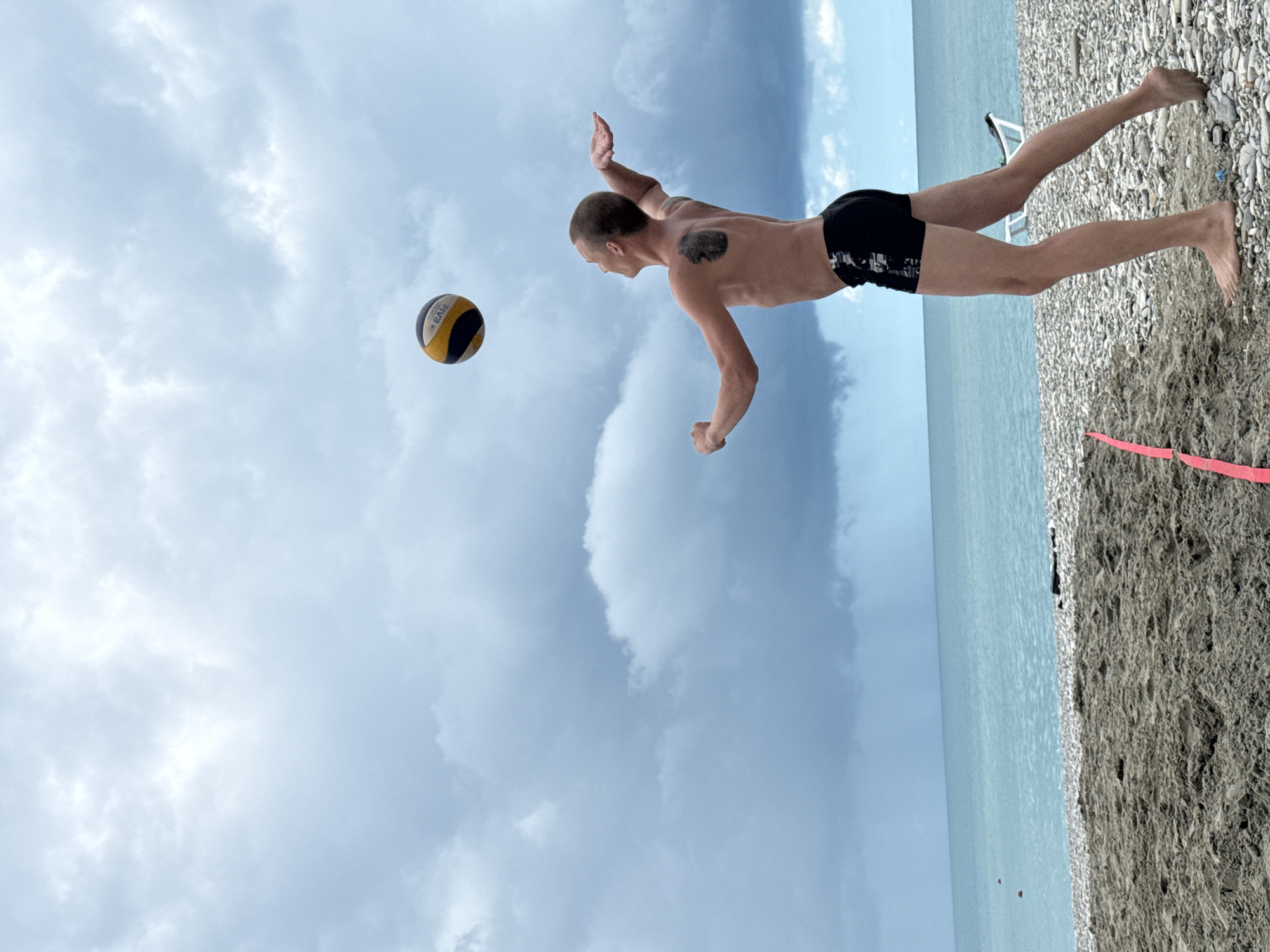 In Nebug, Russia, a young man with a tattoo on his back is playing volleyball on a pebble beach. He is wearing black swim trunks and is in the motion of hitting a volleyball with his right hand. The volleyball, with yellow and black panels, is suspended in the air above and to the left of him. The sea, a vibrant turquoise, stretches out behind him under a cloudy, overcast sky. The foreground shows a sandy beach with a pink line marking a volleyball court.