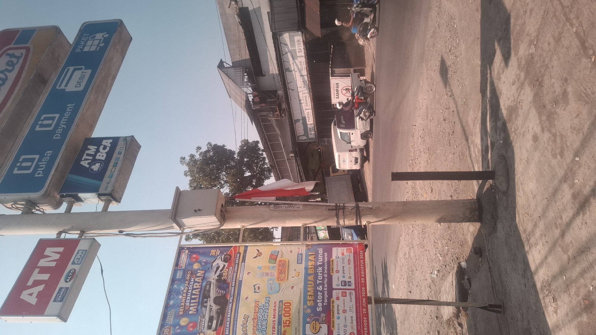 In Rancagoong, Indonesia, a street-level view captures a traffic intersection under a clear sky. Dominating the foreground is a concrete pole supporting several signs. The uppermost sign, red and white, reads "ATM" in large white letters, with smaller text indicating "PRIMA" and "ALTO" below. Adjacent to it is a blue sign with white text, featuring an icon of a plug and the word "pulsa" (top-up/credit), followed by an icon of a square with a dot and "payment," and an icon of a credit card with "DEBIT," and an icon of a truck with "PAKET" (package). Below these, a blue sign displays "ATM BCA" with various card network logos. A red and white Indonesian flag is affixed to the pole, hanging downwards.
To the left of the pole, a large advertisement poster is visible, featuring images of a car, a motorcycle, and cartoon characters, with text such as "BELANJA BERHADIAH MILYARAN" (Shopping with Billion Rupiah Prizes) and "POKÉMON SCHOOL COLLECTION" and "TEBUS MURAH" (Redeem Cheaply). Below this poster, another sign indicates "PERIODE : 25 JUNI 2026 - 05 AGUSTUS 2026" (Period: June 25, 2026 - August 05, 2026).
In the middle ground, vehicles are present. A white van is parked on the right side of the road, next to a grey delivery truck. A person on a black motorcycle, wearing a white helmet, is seen riding from left to right. Further down the road, another person on a lighter-colored motorcycle is also visible. Buildings line the background, with awnings and a sloped roof structure. A sign above a building reads "UD. BERKAH JAYA SAYUR BUAH PARCEL DAGING IKAN TELOR" (UD. BERKAH JAYA Vegetables Fruits Parcel Meat Fish Eggs). Another sign with a red circle and a diagonal line over a silhouette of a person is accompanied by the word "SAMPAH" (Trash). The ground is a mixture of concrete and rough dirt. Sunlight casts shadows across the scene.