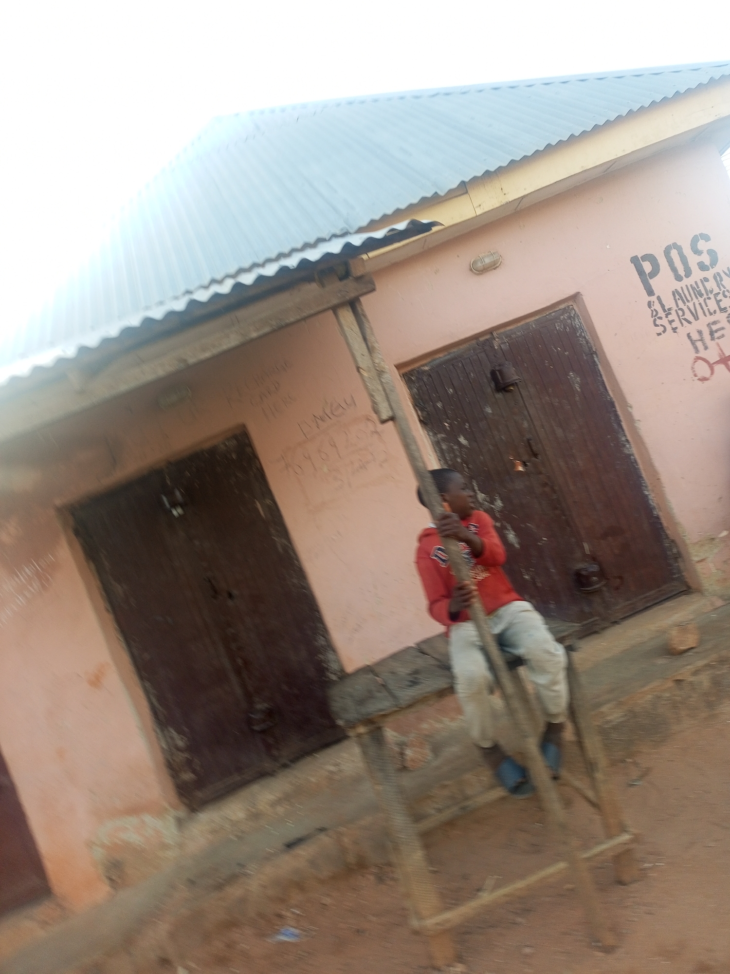 The image shows a young person standing on stilts outside a building in an unknown location. The building is a peach color with two dark brown doors. The roof is made of corrugated metal. Writing is visible on the building walls. A sign on the building reads "POS LAUNDRY SERVICES" with additional text below. The young person is wearing a red shirt, light pants, and blue shoes. The stilts are made of wood. The ground is dirt.