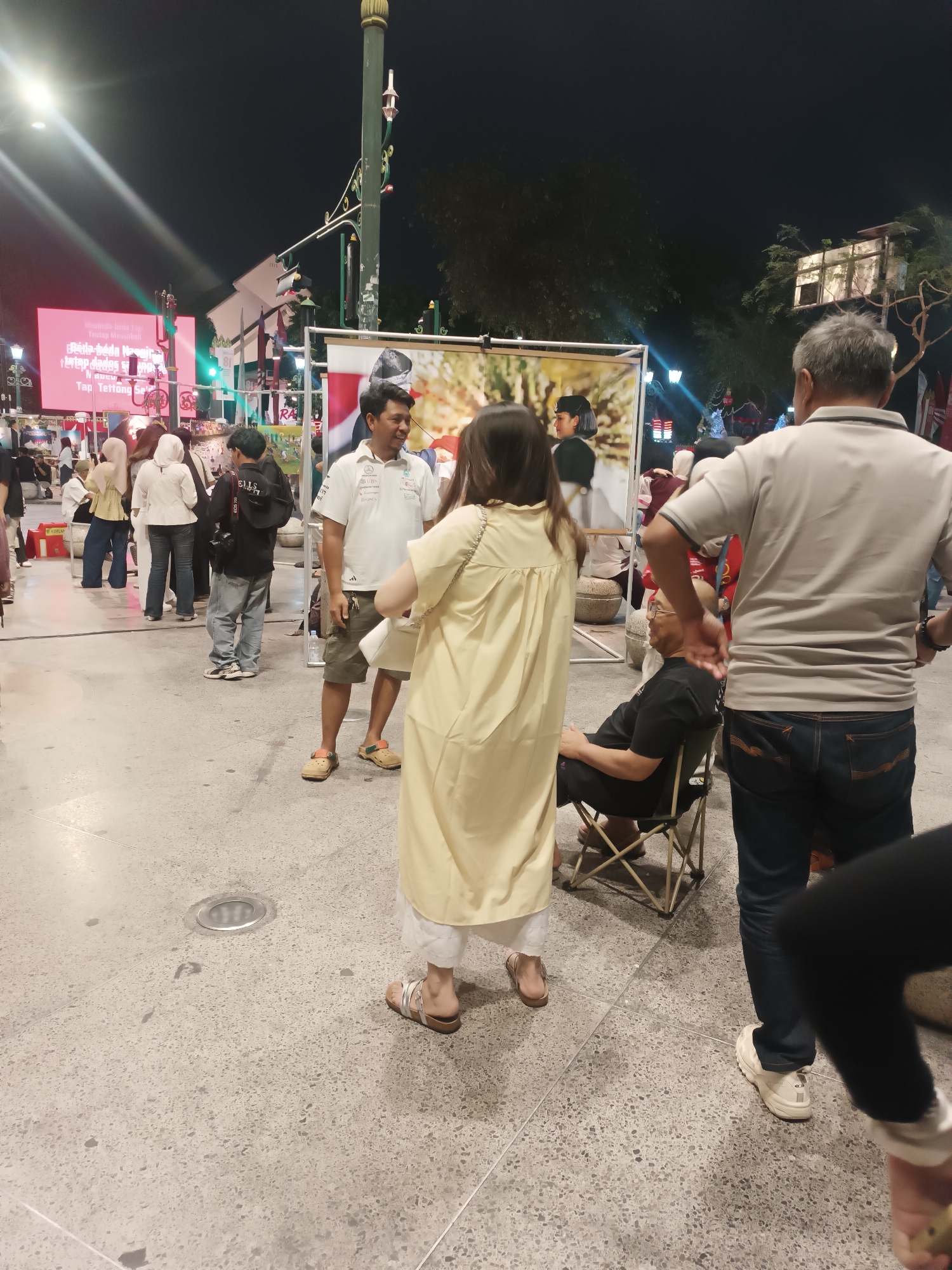 An outdoor nighttime scene captures a group of people gathered around a large poster. In the foreground, a woman in a long, pale yellow dress stands with her back to the camera, facing a man in a white polo shirt and khaki shorts. He is looking to his right and smiling, with keys hanging from his pocket. To the left of the man, a person in a black shirt and jeans with a camera bag over their shoulder is visible. To the right of the man, an older man in a gray polo shirt and dark jeans sits in a folding chair, facing left. In the background, a large pink billboard displays text in an unknown language. A large poster featuring a man's portrait is propped up on a stand. Other individuals are milling about in the middle ground, some wearing headscarves. The ground is paved with light-colored, speckled concrete. Streetlights and other artificial lights illuminate the scene.