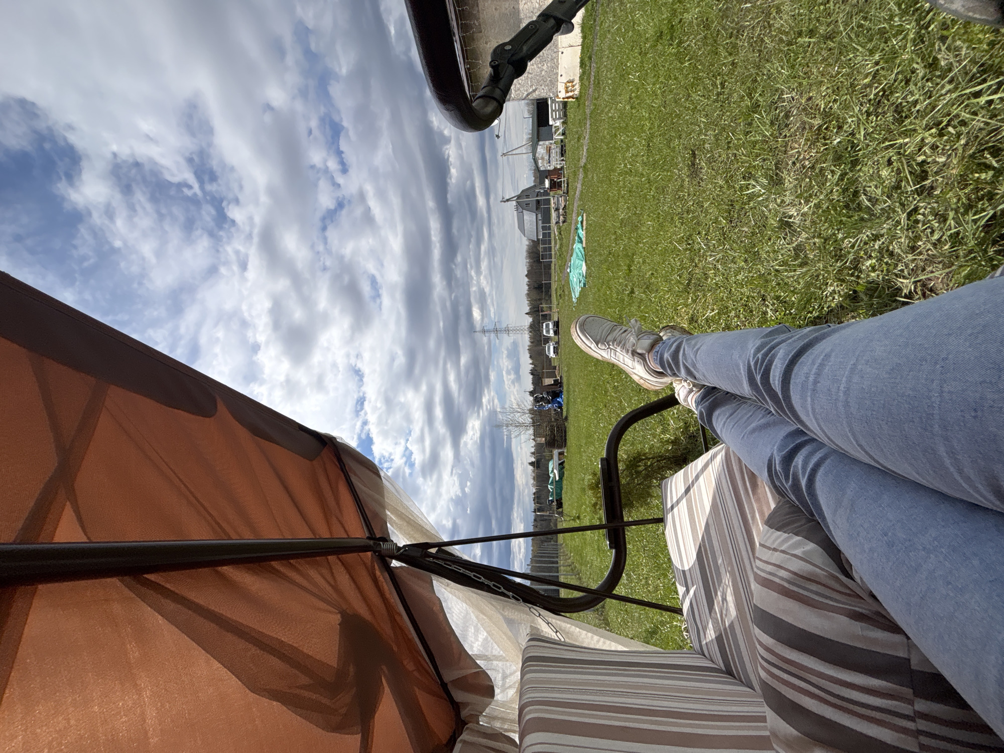 A ground-level, outdoor perspective, possibly from a low-lying chair, presents a partly cloudy sky and a person’s legs stretched out, wearing light blue jeans and grey sneakers. The individual is likely sitting under an orange canopy, supported by black metal rods, which fills the lower left quadrant. The seat appears to be striped and upholstered.
Beyond the canopy and the individual's legs, there's a stretch of green grass. In the background, discernible features include utility poles, several parked cars, and structures that indicate a residential or semi-urban setting, all under the partly clouded sky. The location may be Razdolye, Russia, given the prompt.