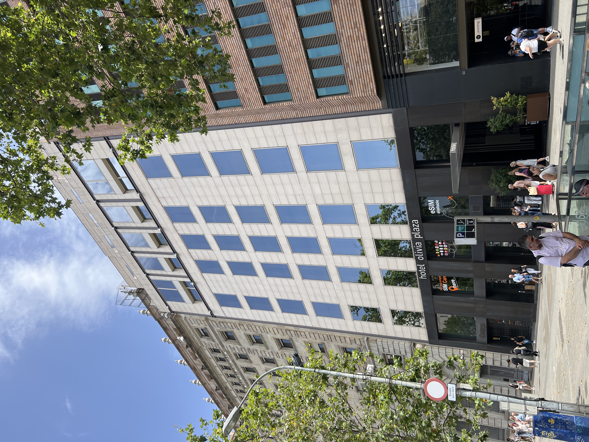 The image captures a street view of the Hotel Olivia Plaza in Barcelona, Spain. The hotel is a modern building with a light-colored stone facade and numerous large, blue-tinted windows. To the left of the hotel is an older, ornate building with a light brown facade and smaller windows. To the right of the hotel, there is a darker brick building with horizontal band windows. Green trees with dense foliage line the street, partially obscuring the buildings.
In the foreground, a street scene unfolds with several people walking on the sidewalk. They are dressed in casual summer clothing. There are street signs, including a red and white circular sign with a diagonal red bar, and a street lamp. The ground appears to be paved with stone or concrete. The sky above is bright blue with scattered white clouds.
The hotel entrance is clearly visible, marked by a canopy and the hotel's name, "hotel olivia plaza," in prominent lettering above the entrance. Several advertisements and signs are displayed on the hotel's ground floor, including one for "eSIM available." There are also reflections of trees and the sky in the hotel's windows. The overall impression is a sunny day in a bustling urban environment.