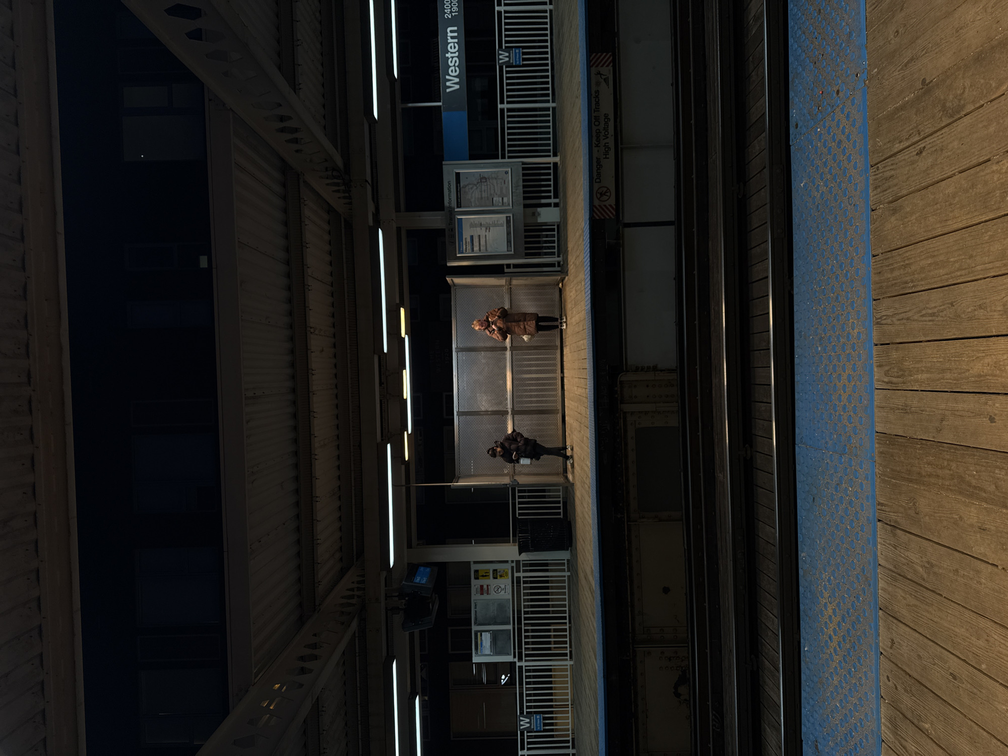 The image depicts a section of the Western CTA "L" station in Chicago, Illinois. Two people are standing on the platform, separated by a gated area. The station signage is visible, identifying it as the "Western" stop. The platform edge is marked with a blue safety strip. The "L" tracks are visible, running parallel to the platform. Overhead, the station structure's metal framework is illuminated by fluorescent lights. The overall scene has a dimly lit, nighttime ambiance.