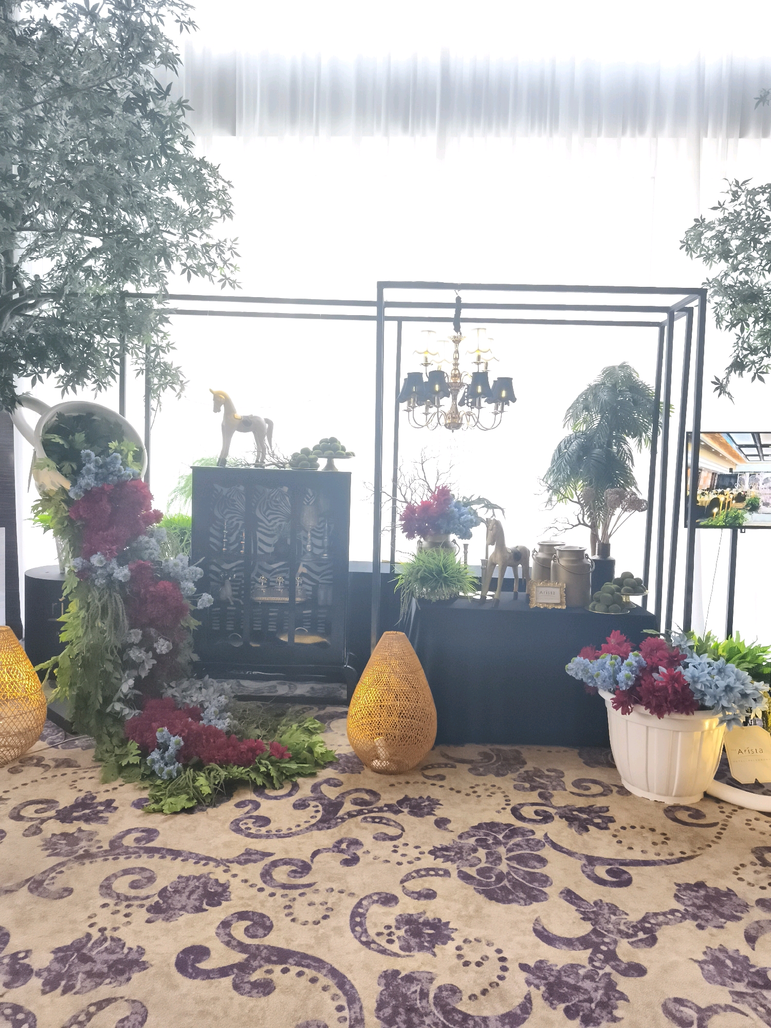The image captures a decorative setup at the Arista Hotel in Palembang, Indonesia. The scene features a display of floral arrangements, decorative objects, and artificial greenery. Prominently displayed is a large white pitcher overflowing with cascading greenery and clusters of red and blue flowers. Beside this, a dark cabinet with a zebra-like pattern stands, topped with a golden toy horse. Further to the right, on a black draped table, are more decorative elements including two metallic milk cans, a framed sign that reads "Arista," a small golden horse figurine, and a tiered stand with green spherical objects. Behind the table, a tall potted palm tree and dried floral arrangements are visible within a black metal frame that also holds a chandelier with black lampshades. The floor is covered with a patterned carpet in shades of beige and purple. Two woven, teardrop-shaped lamps are positioned on either side of the display, and artificial trees with light green foliage frame the left and right edges of the image. A large window with sheer white curtains is in the background, letting in bright light. A digital display screen showing an event setting is visible on the far right. No people are depicted in the image.