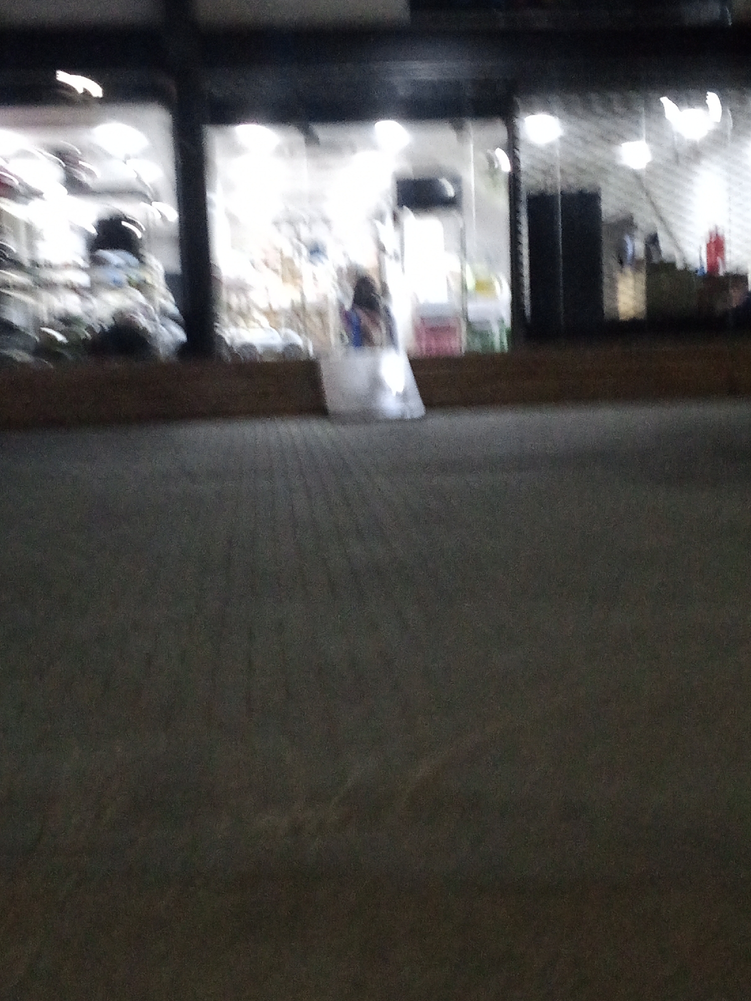 The image depicts a blurred storefront in Ado, Nigeria, viewed from across a paved surface. The store appears to be brightly lit, with merchandise visible through the windows. To the right side of the store is a rolled down corrugated roller shutter door. A translucent plastic bag is located near the store's entrance. The overall scene is dimly lit, with the pavement in the foreground taking up a significant portion of the frame. The blurry nature of the image obscures details.