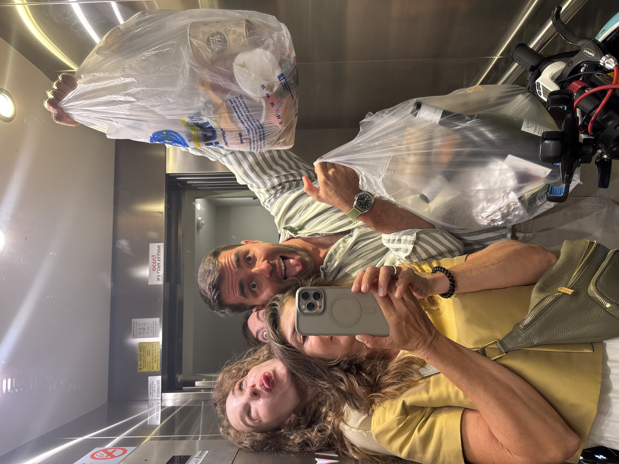 Four individuals are captured in a mirror selfie taken inside a stainless steel elevator in Tel-Aviv, Israel. The elevator features reflective stainless steel walls, recessed overhead lighting, and various informational signs with Hebrew text. One sign on the left displays a "no smoking" symbol. Another sign, partially visible, contains Hebrew text and numbers "000801" and "10800". A distinct red sign near the top right, also reflected, reads "occupi!" in Hebrew with additional Hebrew text.
In the foreground, a woman in her 40s-50s with long, wavy brown hair, wearing a mustard yellow top and a light-colored skirt or pants, holds a grey iPhone 14 Pro/Pro Max with a visible MagSafe ring to take the selfie. She wears a dark beaded bracelet on her right wrist and a ring on her left ring finger. A grey textured fanny pack with gold hardware is slung across her body.
To her left, partially behind her, a woman in her 30s-40s with voluminous wavy brown hair makes a humorous pouting face, looking upwards with her tongue slightly out. She wears a light-colored top.
To the right of the central woman, a man in his 30s-40s with short dark hair and a beard grins broadly, sticking his tongue out and widening his eyes. He wears a light green and white striped short-sleeved button-up shirt and light-colored pants. He holds two clear plastic bags: one raised higher containing various food items including a white cup and what appear to be produce, and another lower one containing a dark-colored bottle (possibly wine) and other items. On his left wrist, he wears a dark green-strapped wristwatch with a dark face.
Behind the central woman and the man, only the eyes and forehead of a fourth person are visible, also making a wide-eyed, humorous expression.
At the bottom right of the image, parts of a bicycle are visible, including black handlebars, brake levers, a phone mount, and what appears to be a light-colored frame with red cabling, positioned near the man's lower plastic bag.