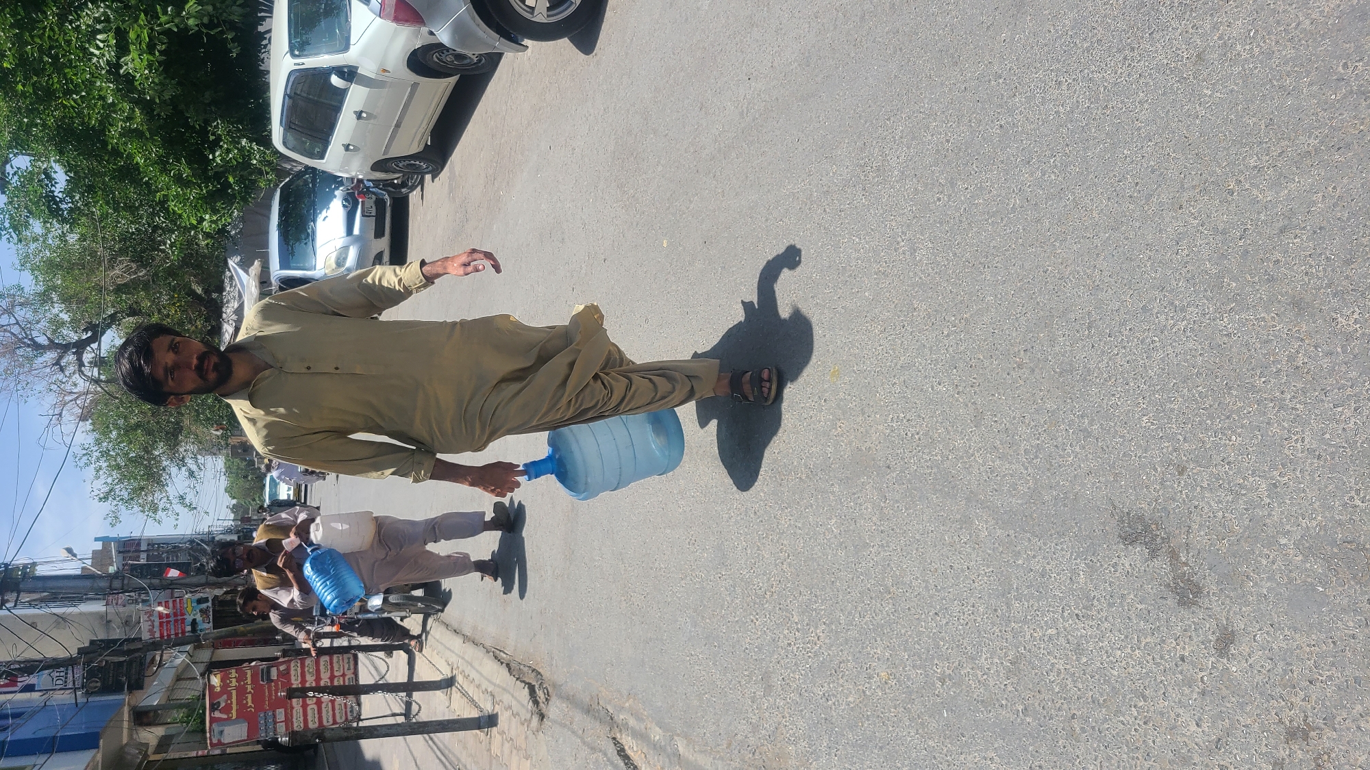 A street scene in Quetta City Tehsil, Pakistan, depicts a man walking on a gray asphalt street, carrying a large blue water bottle. He is wearing a light brown outfit and sandals. The background shows a street with buildings, utility poles, and parked cars. Other individuals are seen on motorcycles on the left side of the image. A tree line is visible in the background. The lighting suggests it is daytime.