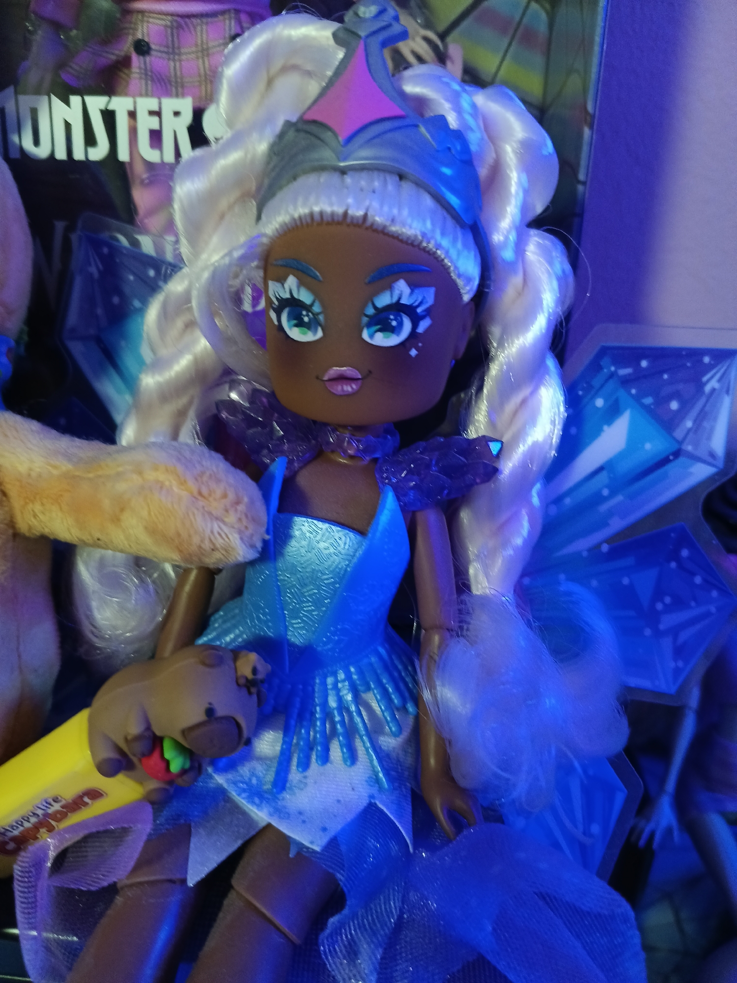 The image is a close-up shot of a doll with dark skin and long, wavy blonde hair. The doll is wearing a light blue, shimmery dress with a flared skirt and a bejeweled collar. It has large, expressive blue eyes with prominent eyelashes and star-shaped glitter accents on its cheeks. The doll's hair is styled with a blue and pink crown-like accessory.
In its right hand, the doll is holding a yellow object that appears to be a candy bar with the text "Happy life! Candy bar" visible. The doll is also holding a small, brown accessory that resembles a stylized creature, with its mouth near a red and green object, possibly representing food.
The background is out of focus but shows what appears to be other dolls or toys, with elements of purple plaid and geometric blue shapes. The lighting is predominantly blue and purple, suggesting an artificial lighting environment, possibly with LED lights.
There is no information in the image that suggests a specific location in the city of Wels, Austria. The image is a studio shot or a close-up of toys.