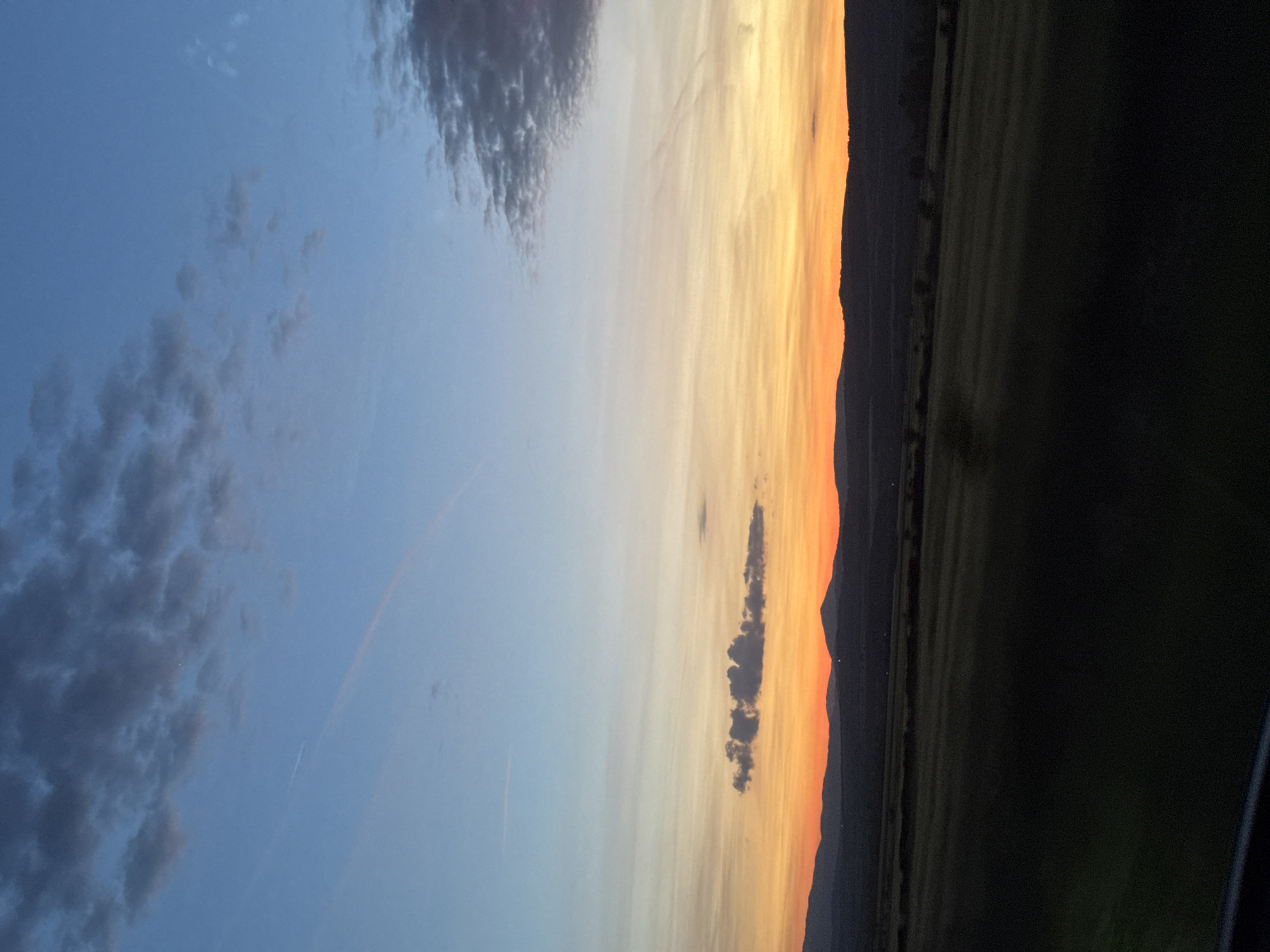 The image captures a dusk or dawn scene in Yahinovo, Bulgaria. The left side of the frame features a blue sky with cumulus clouds. Towards the center, the sky transitions to orange and yellow hues. On the right, dark silhouettes of trees and mountains are visible against the colorful sky. The foreground is obscured by darkness, possibly vegetation or a field.