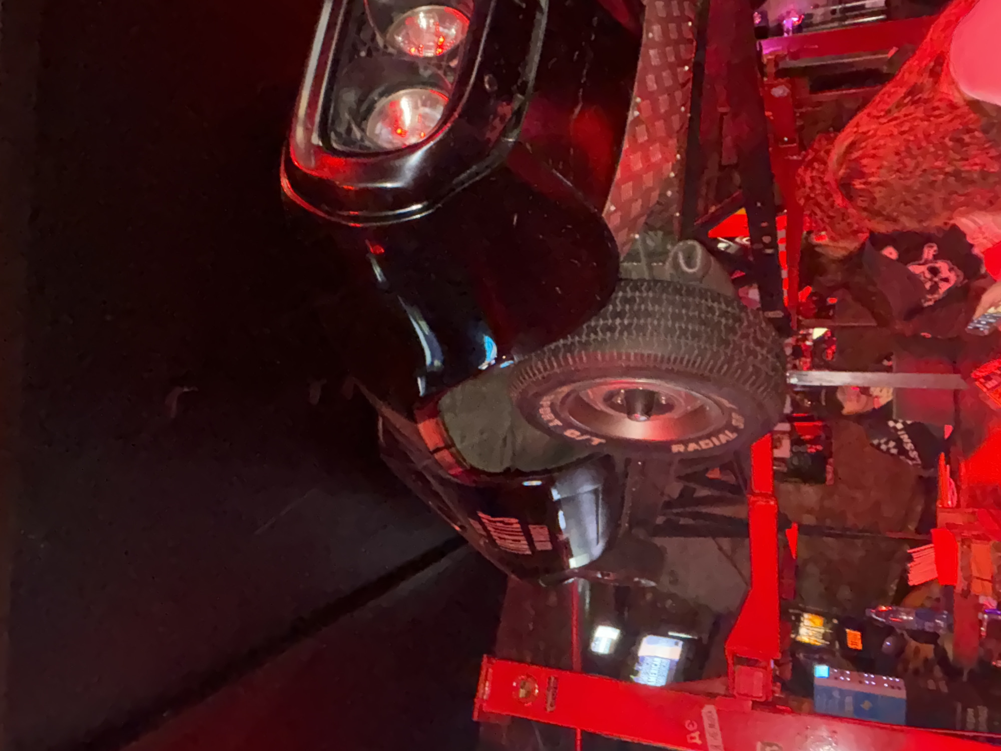 Captured in Saint Petersburg, Russia, the image presents an indoor setting, presumably a bar or establishment with a distinct automotive theme. A black vehicle, possibly a vintage car or a modified model, dominates the scene. Its front end, including the headlights and part of the tire, is prominently displayed. The vehicle is situated on a red platform or stand, possibly for display or decorative purposes.
The surrounding environment features elements consistent with a bar setting. Lights and possibly electronic equipment are visible in the background, contributing to the ambiance. The presence of people is noted, although they appear partially obscured and in the background. The lighting in the area is dynamic, with red hues dominating. The image offers a glimpse into a themed entertainment venue, capturing the unique character of its automotive decor and the bustling atmosphere of Saint Petersburg's nightlife scene.