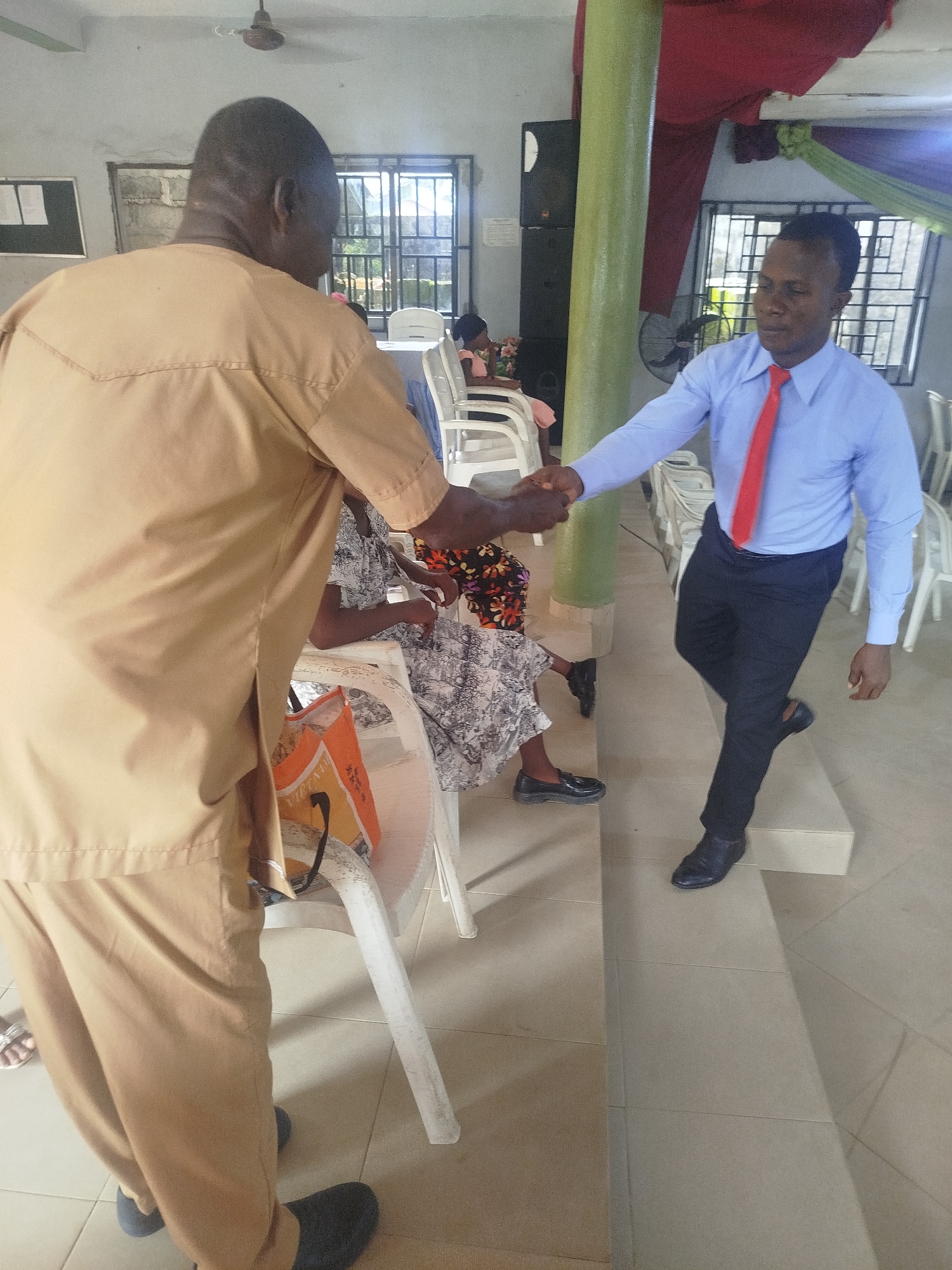 In Ikot Mbonde, Nigeria, a man in a tan two-piece outfit shakes hands with a man wearing a blue shirt and red tie. Both men wear dark shoes. The man in the blue shirt steps up onto a tile-covered platform. Between the men, a woman in a patterned dress sits on a white plastic chair next to a white table holding an orange bag. Other white plastic chairs are visible in the background, near windows with security bars. A green-painted structural column stands near the man in the blue shirt. The scene appears to be indoors, possibly a church or meeting hall. A ceiling fan is visible above the man in tan. A bulletin board is visible on the wall.