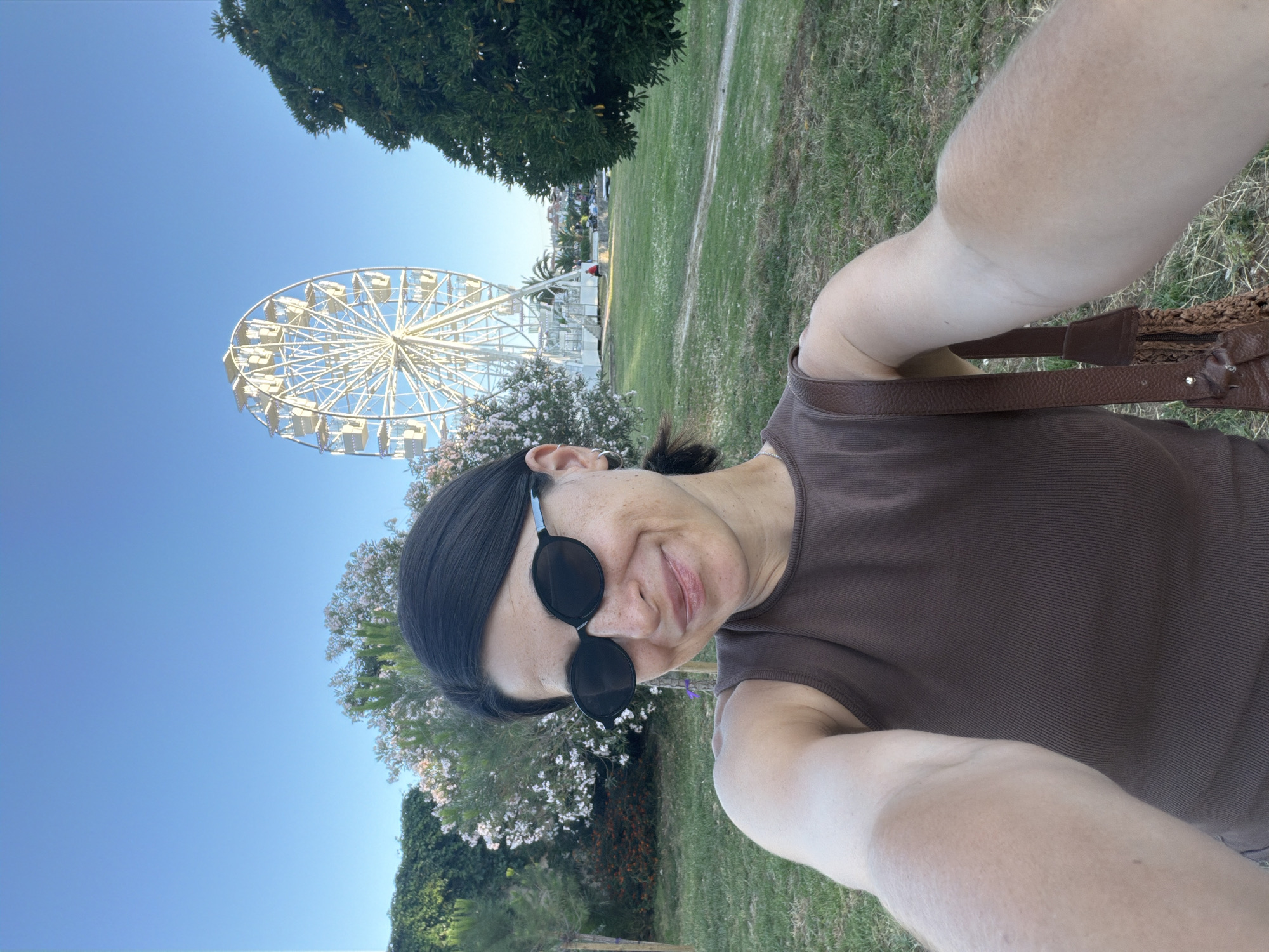 The image displays a selfie of a woman in Budva, Montenegro, against a background featuring a Ferris wheel. The woman, positioned in the foreground, has dark hair pulled back and wears dark oval sunglasses, a brown ribbed tank top, and a silver hoop earring in her visible left ear. A brown leather bag strap crosses her right shoulder. Her face shows visible freckles and a slight smile. The background is dominated by a clear, bright blue sky. A large white Ferris wheel with numerous open gondolas is centrally located in the mid-ground. To the left of the frame, there are green hedges and bushes with white and pinkish flowers. To the right, a large, dense green tree is present. A grassy area extends from the foreground into the mid-ground. Below and behind the Ferris wheel, a white platform structure is visible, with several individuals gathered on and around it, suggesting an active area.