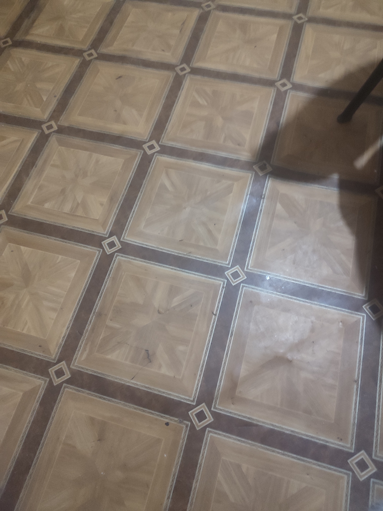 The image shows a tiled floor with a parquet pattern. The tiles are square and arranged in a grid with darker brown grout lines. Each tile features a repeating diamond pattern in a lighter wood tone. There are small, empty square inlays at the intersections of the grout lines. A shadow of an object, possibly a tripod or lamp, is visible on the floor to the right. The floor appears to be in an indoor setting, likely a room in Chernivtsi, Ukraine.