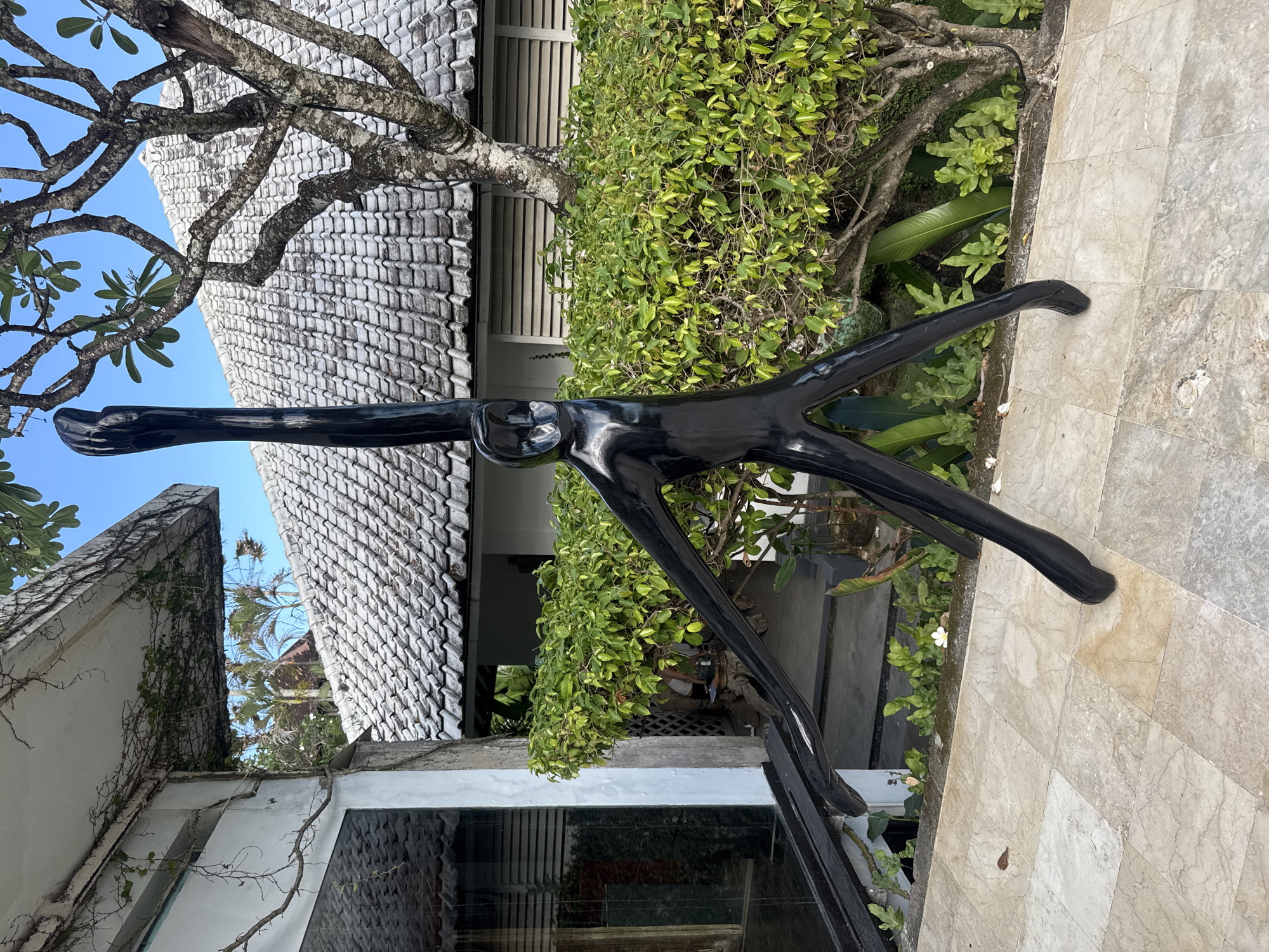 A black metal sculpture resembling a stretched human figure is centrally positioned outdoors in North Kuta, Indonesia. The sculpture stands on a tiled stone surface, partially surrounded by greenery. Behind the sculpture, there is a building with a white tiled roof and white exterior walls, showing a blend of modern and traditional architectural styles. A tree with bare branches is present in the upper left corner, partially obscuring the sky. The building also features reflective glass panels that reflect the surrounding environment, including palm trees.