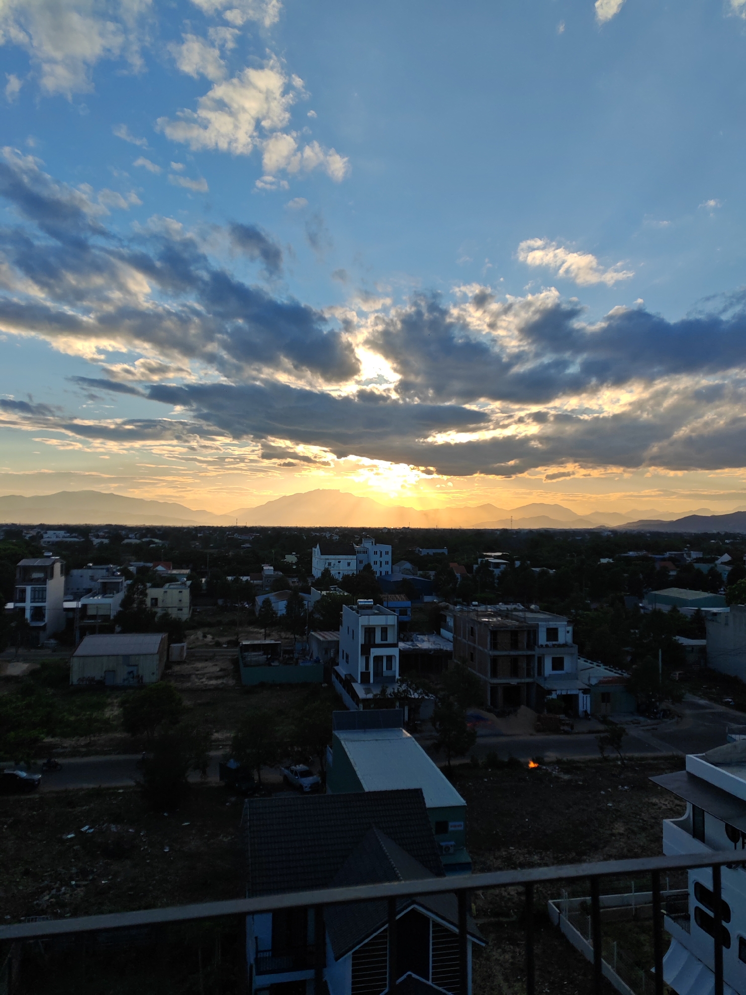 This is a high-angle, medium shot of a residential area in Đà Nẵng, Vietnam, during a sunset. The sky is a dynamic mix of blue and grey clouds, with rays of sunlight breaking through the clouds and illuminating the distant mountains. The sun is setting behind the mountains, casting a warm golden glow across the landscape.
In the foreground, the edge of a balcony railing is visible. Below, a scattered collection of buildings, some under construction and others seemingly completed, are interspersed with trees and vegetation. Roads are visible, with a few vehicles, including a car and a motorcycle, present. There is evidence of recent construction activity, with piles of building materials such as sand visible in some areas. A small, bright orange fire is visible in the middle ground, potentially a small controlled burn or waste disposal. The overall impression is of a developing urban fringe bathed in the dramatic light of sunset.