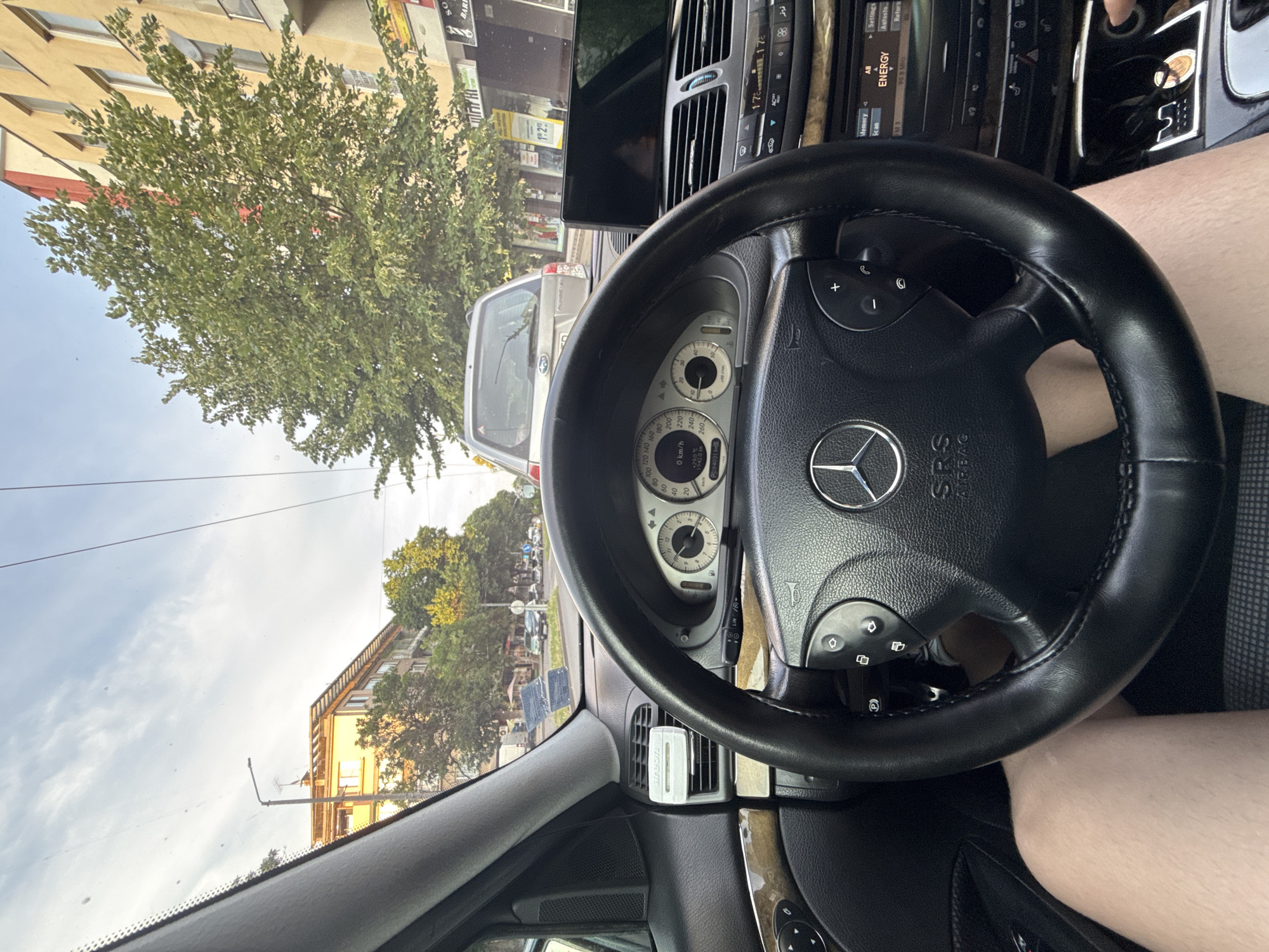 The image captures the interior of a Mercedes-Benz vehicle, likely in Stara Zagora, Bulgaria, viewed from the driver's perspective. The steering wheel, featuring the Mercedes-Benz logo, is prominent in the foreground. The dashboard displays the car's instrumentation, including a speedometer and tachometer, with the car appearing to be stationary at 0 km/h. The central console shows climate control settings and an infotainment screen. Outside the car, a street scene is visible, with other vehicles, buildings, and trees. A silver Subaru Forester is directly in front, indicating traffic. The lighting suggests it is daytime, possibly late afternoon, with a partly cloudy sky. The car's interior shows signs of use, with a air freshener clipped to the vent. A person's legs are visible in the lower part of the image, suggesting the driver. The environment appears to be an urban setting, consistent with a city like Stara Zagora.