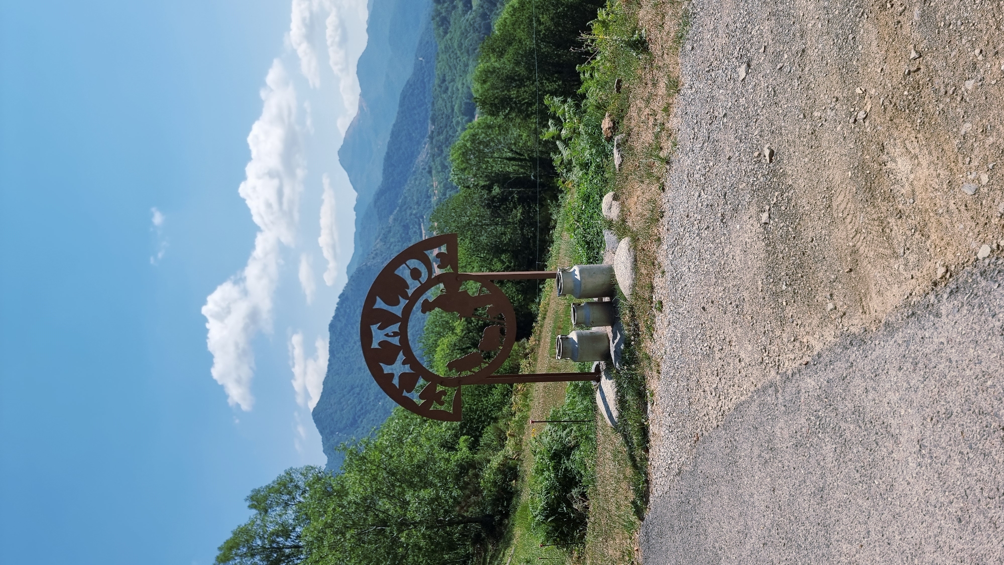 A rustic metal sculpture, featuring a silhouette of a farmer with sheep, is positioned in front of a backdrop of lush green mountains under a clear blue sky with scattered white clouds. Three vintage milk cans are placed at the base of the sculpture, resting on rocks. The scene is located on a grassy area with a gravel and asphalt path in the foreground. The sculpture's arch is decorated with cutout stars. The overall setting suggests a rural landscape in or near Camprodon, Spain, possibly a farm or a scenic viewpoint.