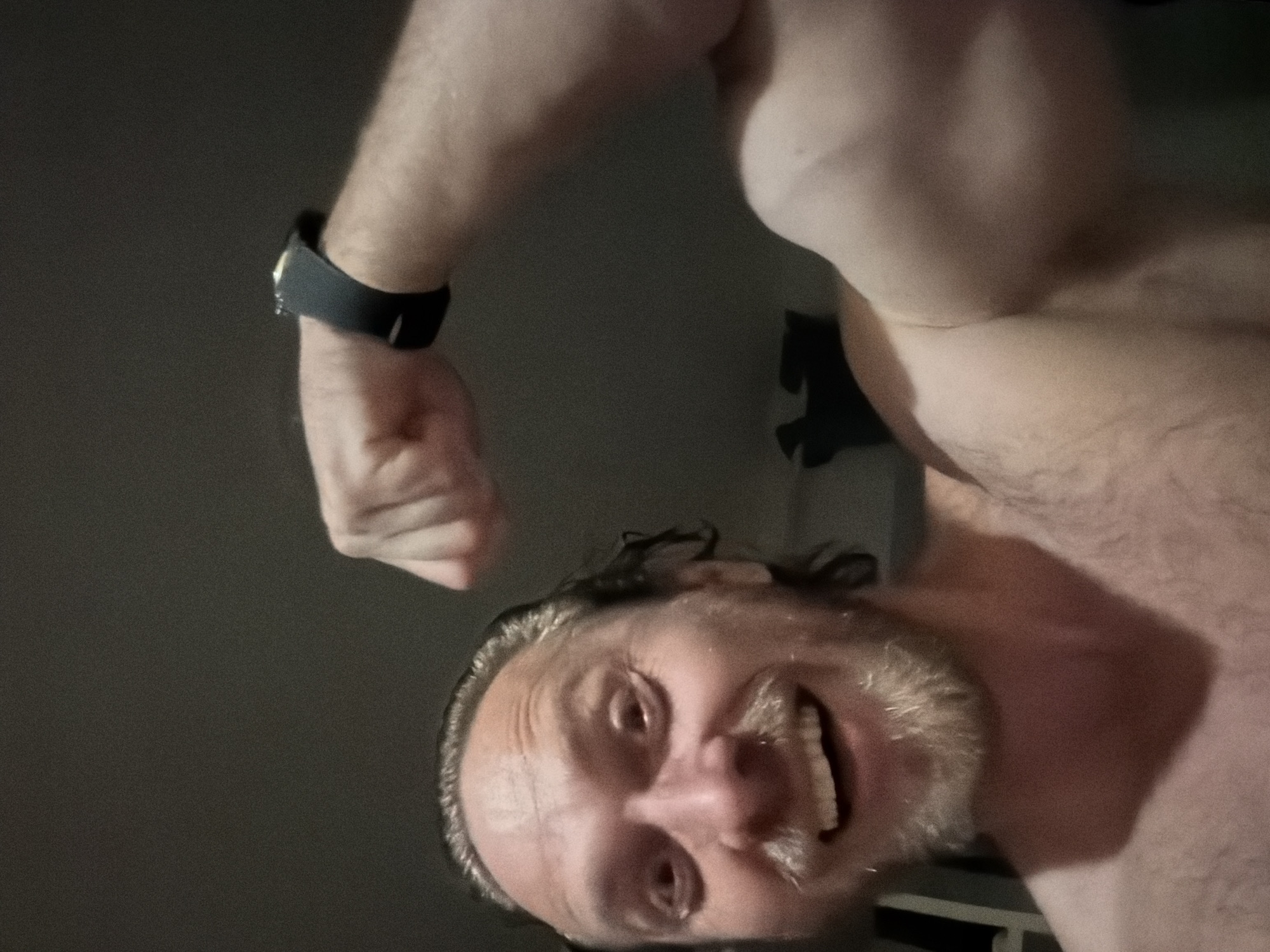 An adult male is depicted indoors in Saint Petersburg, Russia. The man has fair skin, wet or slicked-back dark blonde hair, and a light-colored beard and mustache. His eyes are wide, and his mouth is open in a broad smile, revealing white teeth. He is bare-chested, with visible chest hair, and is flexing his left bicep towards the camera, clenching his fist. A dark-colored smartwatch with a black band is worn on his left wrist. The lighting is dim and artificial, casting some shadows. The background is dark, blurry, and indistinct, suggesting a private indoor environment.