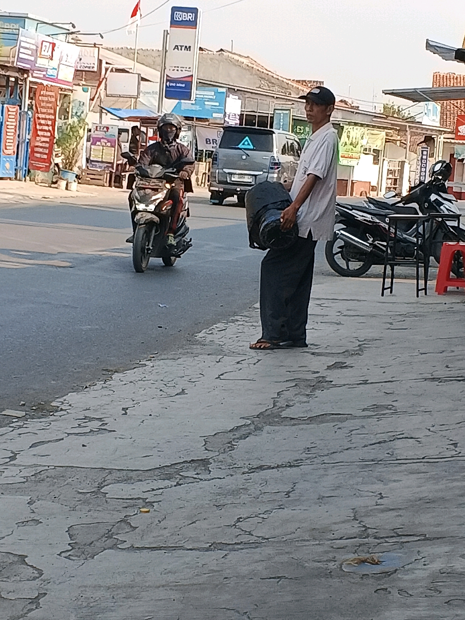 In the city of Taman, Indonesia, a scene unfolds on a street with a sidewalk adjacent to a road. A man, wearing a white short-sleeved shirt, dark pants, a black baseball cap, and flip-flops, stands on the sidewalk holding a large, dark cylindrical object. He appears to be carrying it. Beside him, parked on the sidewalk, is a black motorcycle. In the background, across the road, is a row of shops and buildings with various signs, including a prominent "BRI ATM" sign. Further down the road, a person is riding a motorcycle, wearing a helmet and dark clothing. The sky is bright, suggesting daylight. The sidewalk has a textured, cracked surface.