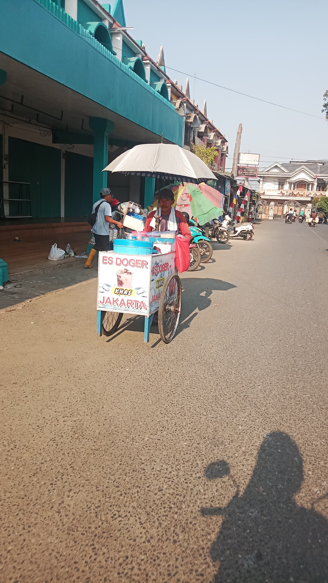 In the city of Jawa, Indonesia, a street vendor is selling ice from a cart on a sunny day. The vendor, a man with a mustache, sits behind the cart, which is decorated with colorful signs advertising "ES DOGER KHAS JAKARTA." A large white umbrella shades the vendor and the cart. In the background, several motorbikes are parked, and buildings line the street. The street is paved with asphalt, and a shadow of a person on a motorbike is cast on the road, indicating the presence of other vehicles. The overall scene depicts a typical street-side marketplace in Indonesia.
