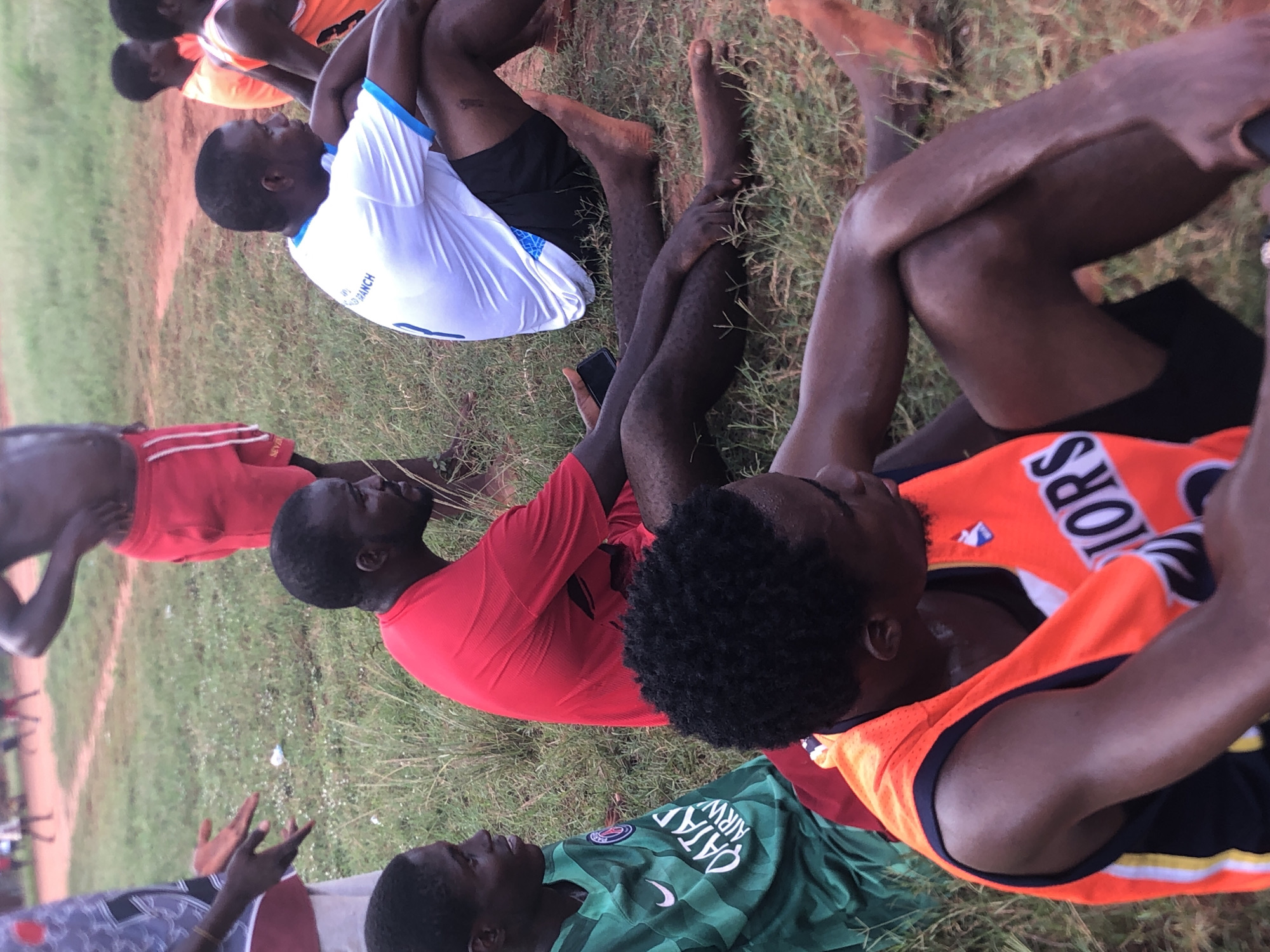 This image captures a group of at least six young to adult men resting on a grassy and dirt field, likely after engaging in physical activity. The setting appears to be an outdoor sports ground in Ukwu-Abwa, Nigeria.
The men are positioned in various states of rest on the ground, mostly lying on their backs or sitting with bent knees. In the foreground right, a man wears an orange and black sleeveless jersey with partial text "S" and "IORS" (likely part of a team name). He has dark, curly hair and is looking upwards. Adjacent to him, another man in a red short-sleeved shirt is also looking skyward. To the left, a man in a white and blue athletic shirt, which reads "WORLD BRANCH", is lying down with his knees bent. Another man, wearing a red short-sleeved top, is centrally located, holding a black smartphone in his hand. Further back and to the left, an individual in a green soccer jersey featuring "QATAR AIRWAYS" and a Nike swoosh is also seen resting.
The ground is a mix of green grass and reddish-brown soil, characteristic of a field used for sports. The lighting is soft and warm, suggesting the time of day is late afternoon or early evening. The men appear relaxed, indicating a period of recovery or casual rest.
