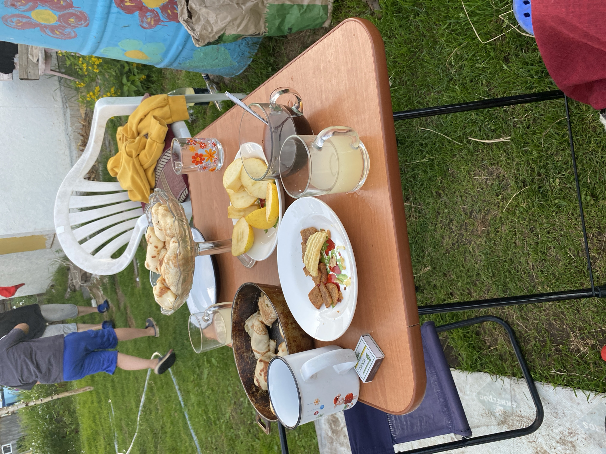 The image depicts an outdoor scene with a folding table set for a meal. On the table are several items, including a bowl of pastries, a frying pan with what appear to be crepes or blinis, a white mug with a cartoon character, a plate with crackers and some sort of spread, and several glasses filled with drinks. In the background, two people are partially visible, appearing to walk away from the camera. One person is wearing blue shorts and a grey shirt, and the other is wearing grey shorts and a dark top. A white plastic chair with a yellow sweatshirt draped over it is also present. The environment is grassy, with some yellow flowers visible. A large blue barrel decorated with colorful flowers is situated to the right of the table. The setting suggests a casual outdoor gathering or meal. The presence of a matchbox on the table implies that smoking might have occurred. The image is taken from a slightly elevated perspective, looking down at the table.
