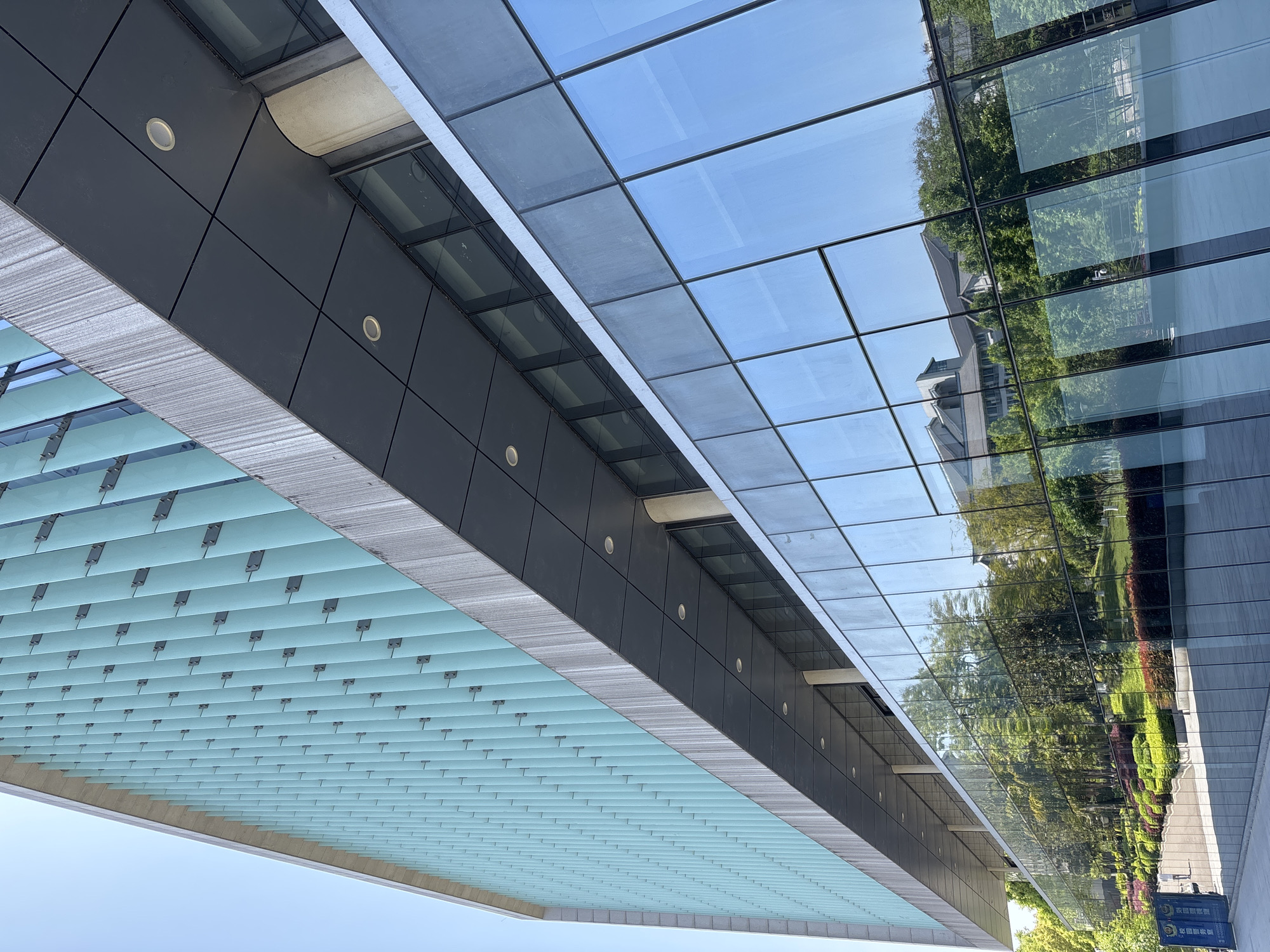 The image shows a modern building in Jiangjin, China. The building's facade is primarily made of glass, reflecting the surrounding trees, sky, and another building. There are dark gray panels with recessed lights running along the building's edge. Below the panels is a blue-toned covering with a grid pattern. The sky is visible in the bottom left corner, and a sign with Chinese characters is on the right side of the image. The photograph appears to be taken from a low angle, looking upward at the building.