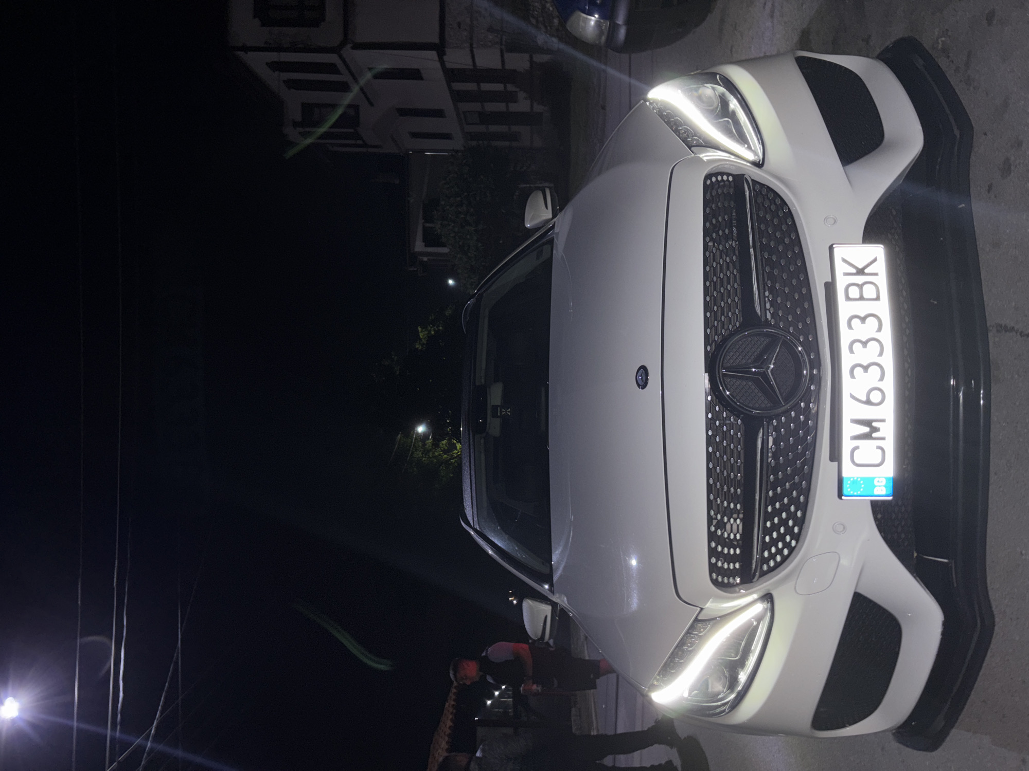 This forensic description details a night scene in Zlatograd, Bulgaria. A white Mercedes-Benz C-Class (W205 generation, likely an AMG-line variant) station wagon is prominently featured, centered and facing the viewer. Its LED headlights are illuminated. The car displays a Bulgarian license plate "CM 6333 BK" with the "BG" EU country code. An aftermarket black front splitter is attached below the bumper.
On the left side of the image, two males are visible. One, dressed in dark clothing, is partially obscured by the car's hood. The other, wearing a dark sleeveless vest over a lighter shirt and dark shorts, holds a dark object, possibly a beverage can, and is facing the first individual. Behind them, a building with a dark tiled roof and visible wooden elements can be discerned.
To the right of the Mercedes, the front section of a blue vehicle is partially visible. In the background on the right, a multi-story white building with distinct dark windows and traditional architectural features stands under the low light. Overhead, a bright street light is visible in the upper left corner, casting light, and several power lines traverse the dark sky. Green light trails, possibly lens flares or light reflections, are present in various parts of the upper and middle sections of the image. The ground appears to be a paved surface.