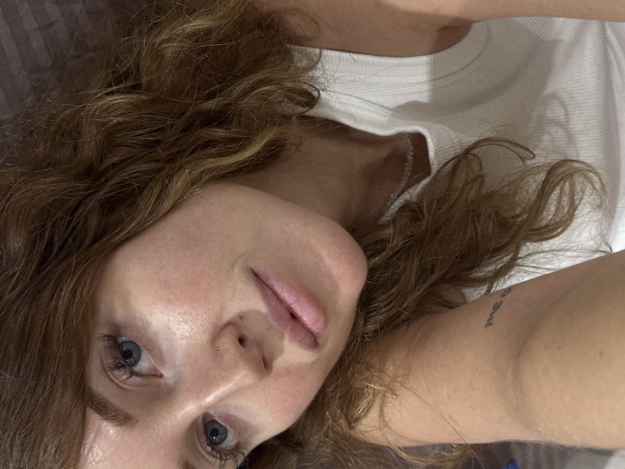 The image is a close-up portrait of a young adult female, positioned at an oblique angle as if lying down. Her face is central, with her head resting to the left and her body extending to the right of the frame. She has long, wavy, light brown or auburn hair that frames her face and spreads around her.
The subject is wearing a white, ribbed top or undershirt. Her blue eyes are open and directed towards the viewer, and her lips are slightly parted. A small, subtle nose stud is visible on her left nostril. Her skin appears fair. On her right upper arm, a small, dark tattoo is visible, which appears to read "me o" or "me e", possibly part of a larger word or phrase.
The setting appears to be indoors, likely a personal space, possibly a bedroom, indicated by a dark, soft-textured background that resembles bedding or a pillow behind her head. The lighting is soft and somewhat dim, suggesting an indoor artificial light source or low natural light. There are no specific location cues visible in the image itself to confirm the provided context of Chaikovsky, Russia. Her expression is calm and neutral.