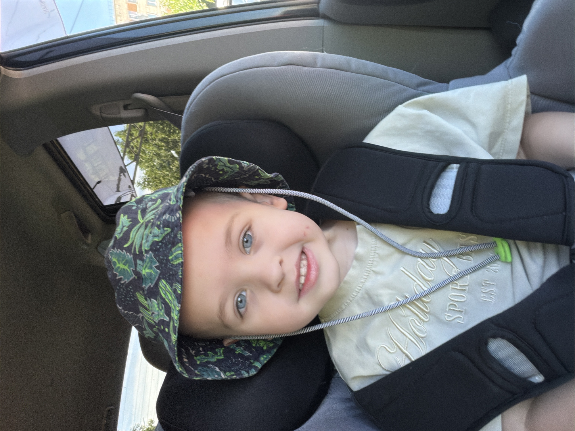 A young boy with blue eyes and light brown hair, wearing a patterned bucket hat and a light-colored t-shirt with embroidered text that reads "Holiday SPORT BITS EST 2004," is sitting in a car seat inside a vehicle. The car appears to be parked on a street in Khabarovsk, Russia, as indicated by the buildings and trees visible through the car window. The sun is shining, casting shadows and illuminating the scene. The boy is looking directly at the camera with a slight smile. The car seat is grey and black, with a black safety harness. The boy's right cheek has a small red mark.