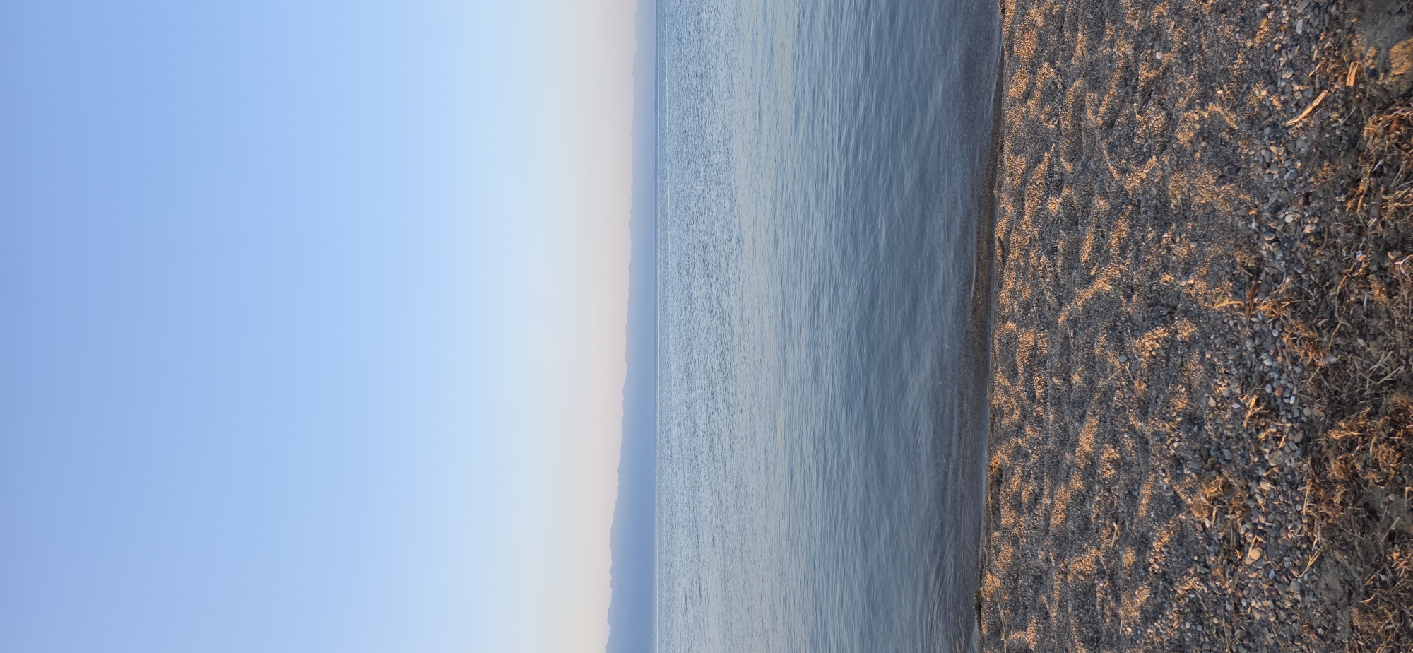 A serene, wide-angle vertical shot captures the tranquil scene at the shores of Issyk-Kul lake, near the village of Boz Beshik, Kyrgyzstan. The image is divided into three horizontal bands: a clear, pale blue sky occupies the top third, transitioning to a soft, hazy horizon line of distant mountains in muted blues and purples. The middle section is dominated by the calm, glassy surface of the lake, reflecting the sky and distant landscape with subtle ripples near the shore. The foreground consists of a pebbly beach, illuminated by warm, low-angled sunlight casting dappled shadows and highlighting the texture of the stones and scattered dry vegetation. There are no people or explicit actions depicted, emphasizing the natural, undisturbed beauty of the environment. The overall atmosphere is one of peacefulness and stillness, typical of early morning or late afternoon light.