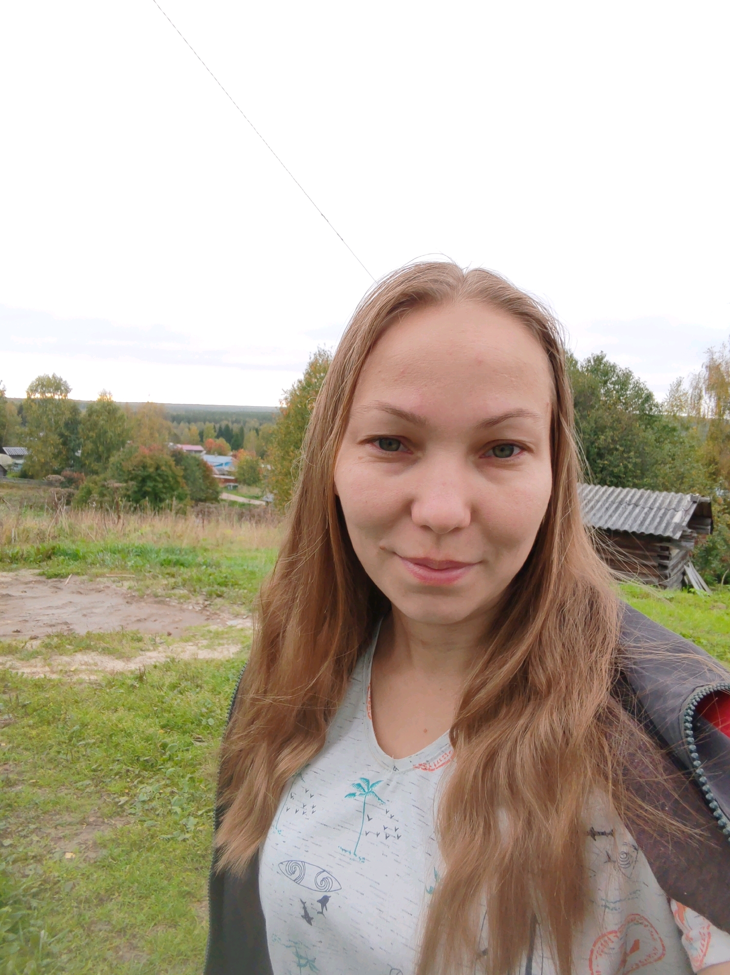 A young woman with long, light brown hair is taking a selfie outdoors in a rural setting. She is wearing a light blue t-shirt with a graphic print and a dark jacket. The background shows a landscape with green grass, a dirt path, trees with autumn foliage, and several small houses and structures. A rustic wooden shed with a corrugated roof is visible to her right. The sky is overcast. The location appears to be a village in the vicinity of Чухлэм, Russia, given the architectural style of the buildings and the natural environment.