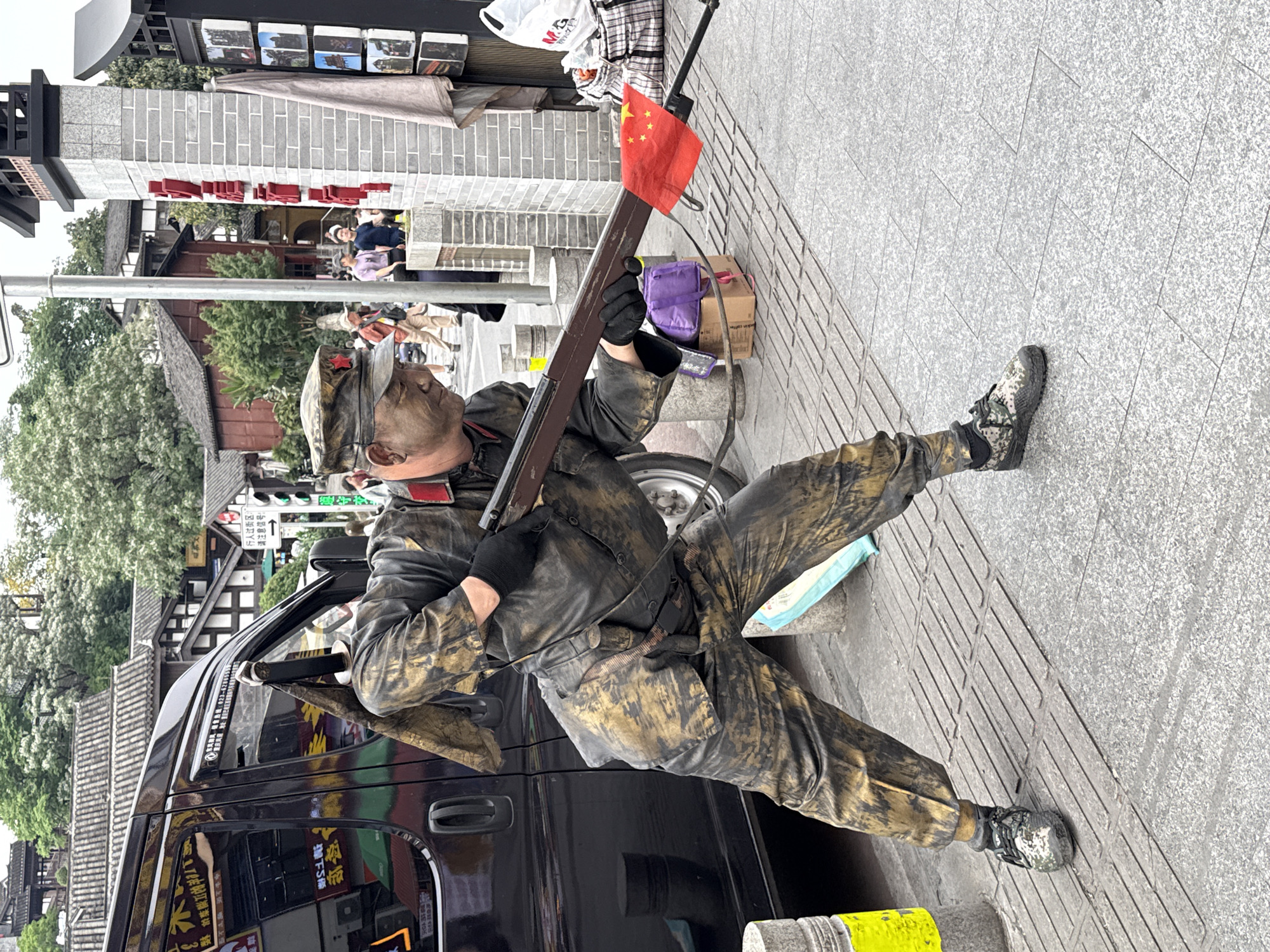 The image shows a man in a camouflage military-style uniform sitting on the ground with a Chinese flag attached to a rifle. He is positioned next to a black van. The background includes buildings with traditional Chinese architecture, suggesting a tourist area. Signage with Chinese characters is visible on the van and nearby buildings. The location is likely in the Shapingba District, China, based on the Chinese signage and architectural style.