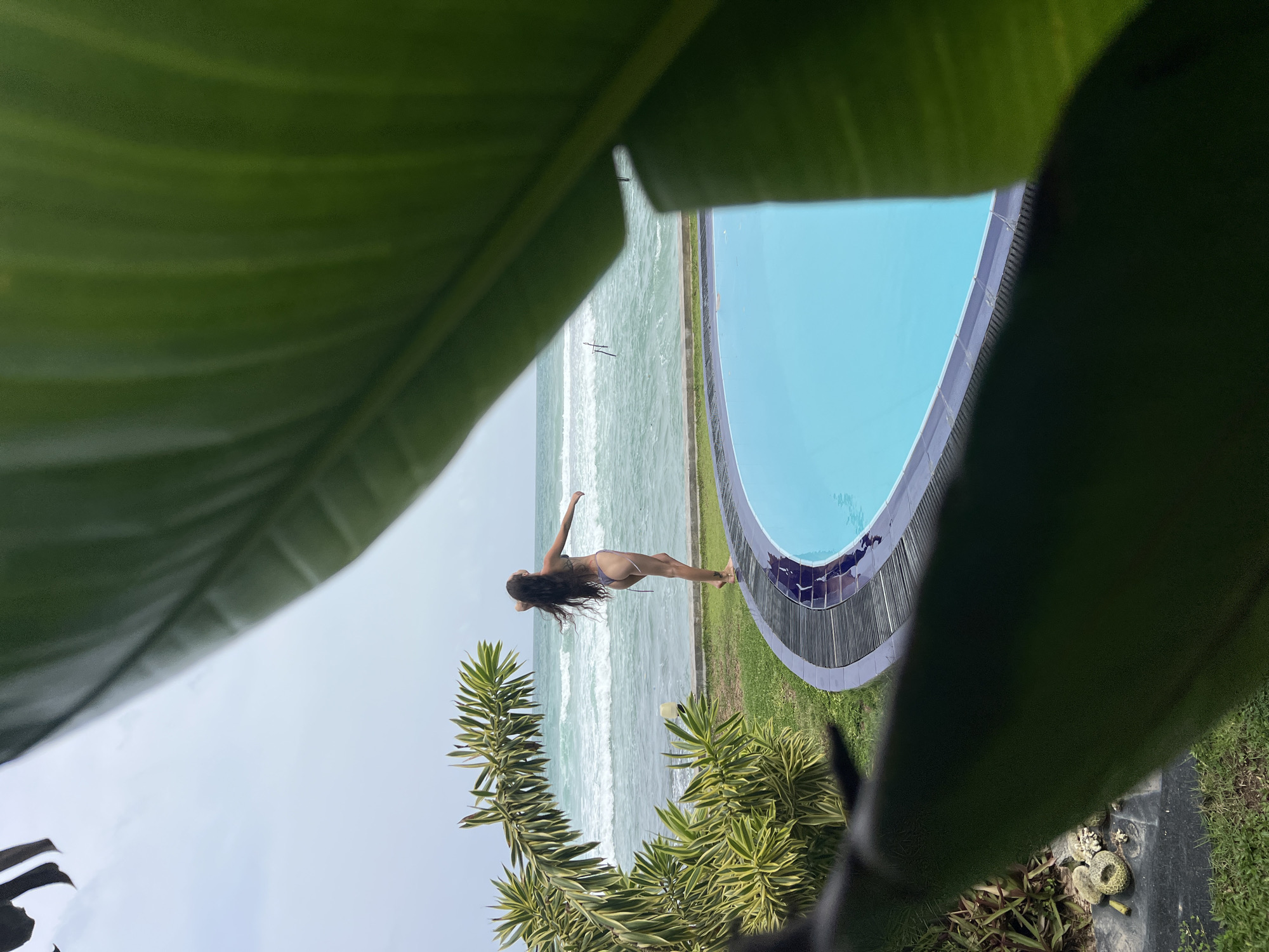 The image depicts a scene in Goviyapana, Sri Lanka, framed by a large green leaf in the foreground. Beyond the leaf, a woman in a swimsuit stands on a grassy area adjacent to a curved swimming pool filled with light blue water. The ocean, marked by breaking waves, stretches out to the horizon beyond the pool. A small tree is to the left.